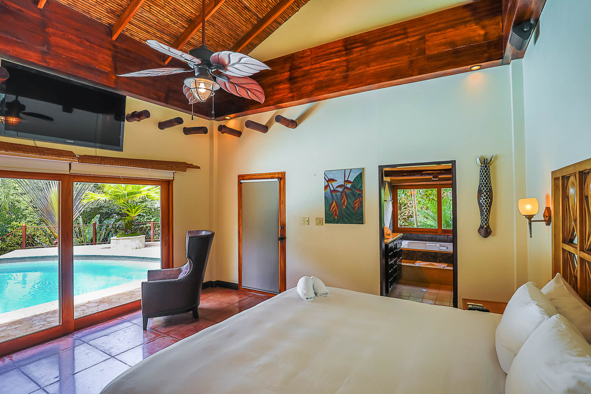 This bedroom offers a view of the adjacent pool and tropical forest through large sliding glass doors, featuring a high wooden beamed ceiling, terracotta floor, and an open doorway to an en-suite bathroom.