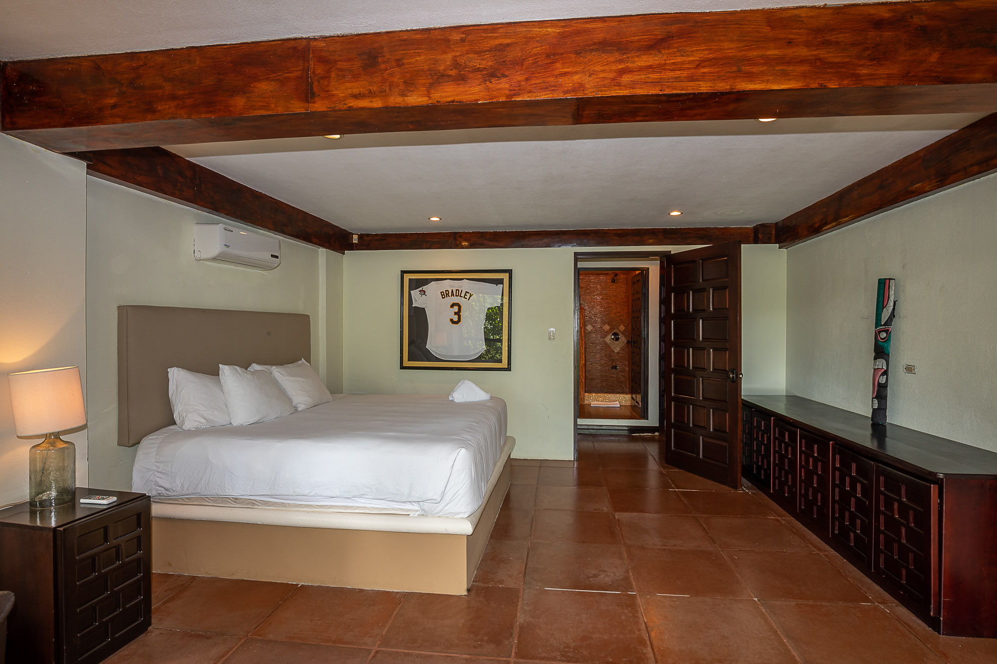 This bedroom features a large bed under exposed wooden ceiling beams, terracotta-style flooring, and an open doorway leading to a private bathroom.