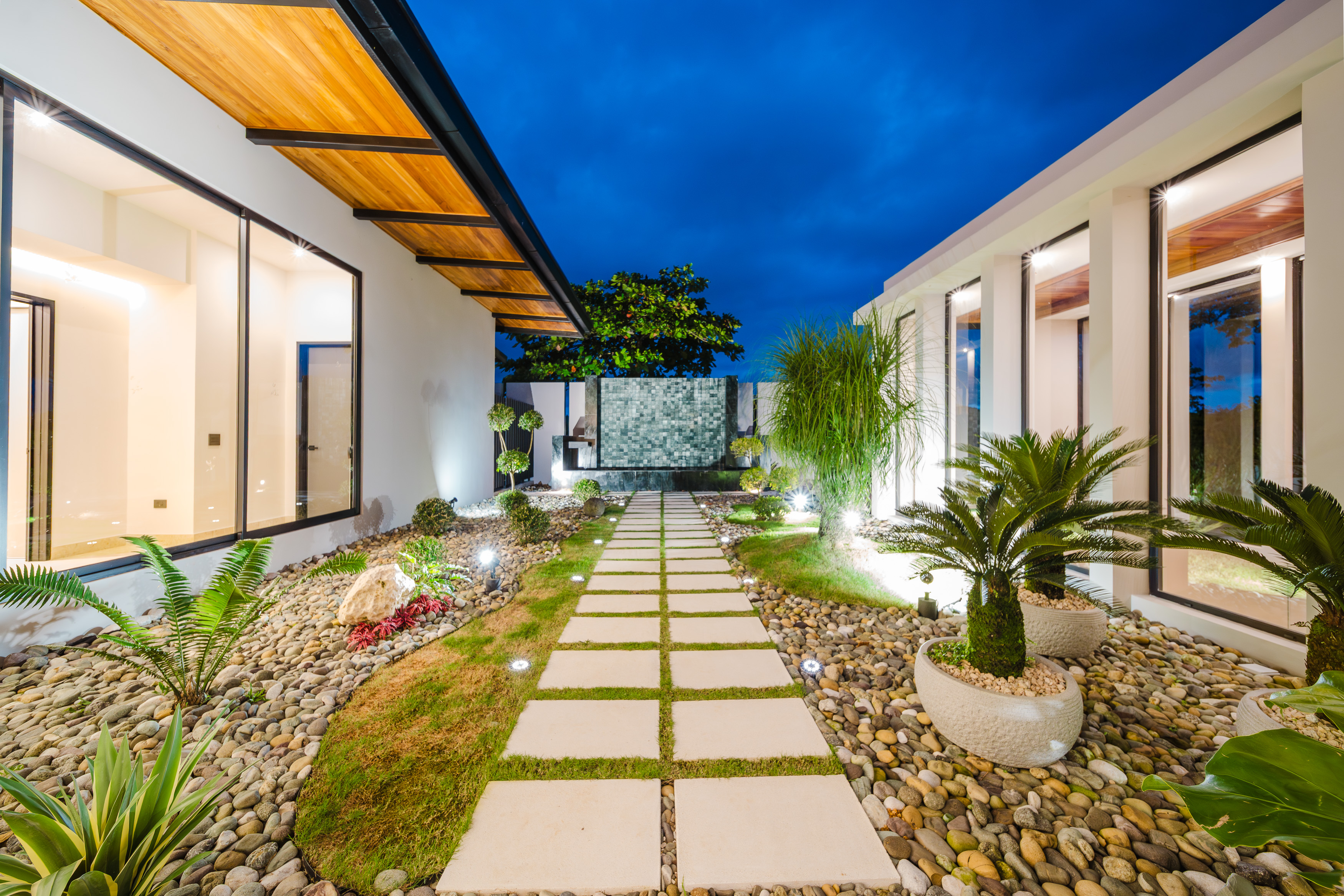A modern villa entrance features an illuminated pathway through tropical gardens, flanked by glass walls revealing warm interiors under a twilight sky.