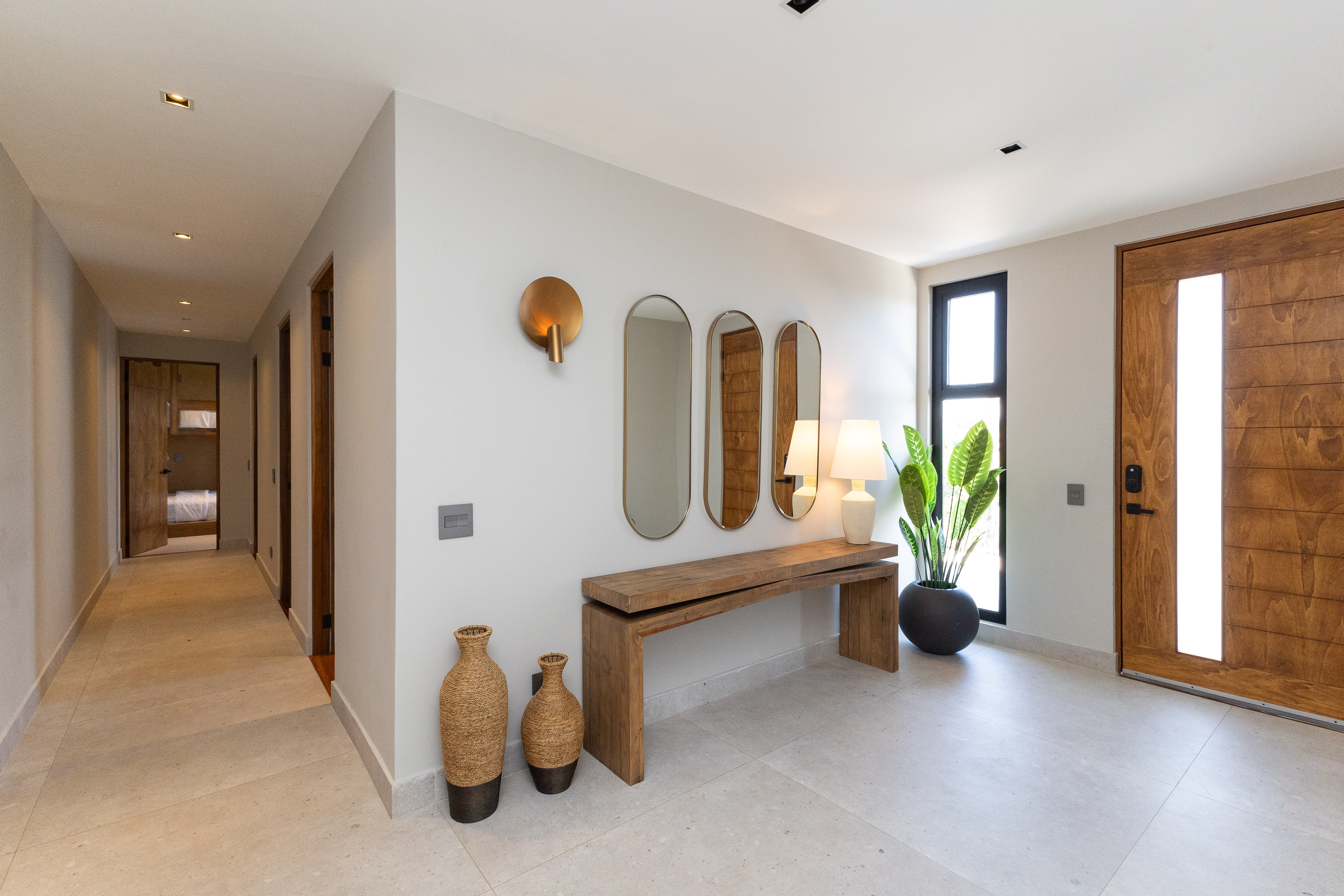 This bright, modern entry area features a large wooden door with a vertical glass panel, leading to a hallway accessing other private rooms within the villa.