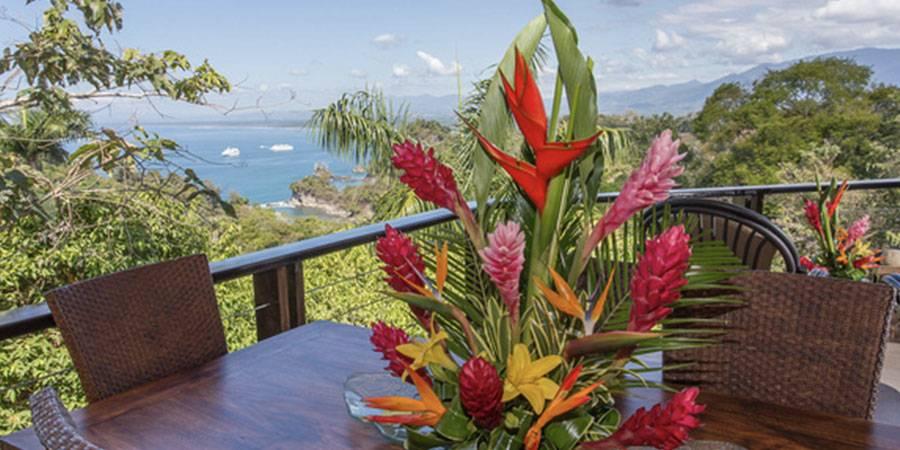 This outdoor terrace with a wooden table and wicker chairs provides a panoramic view of the Pacific Ocean and surrounding tropical rainforest.