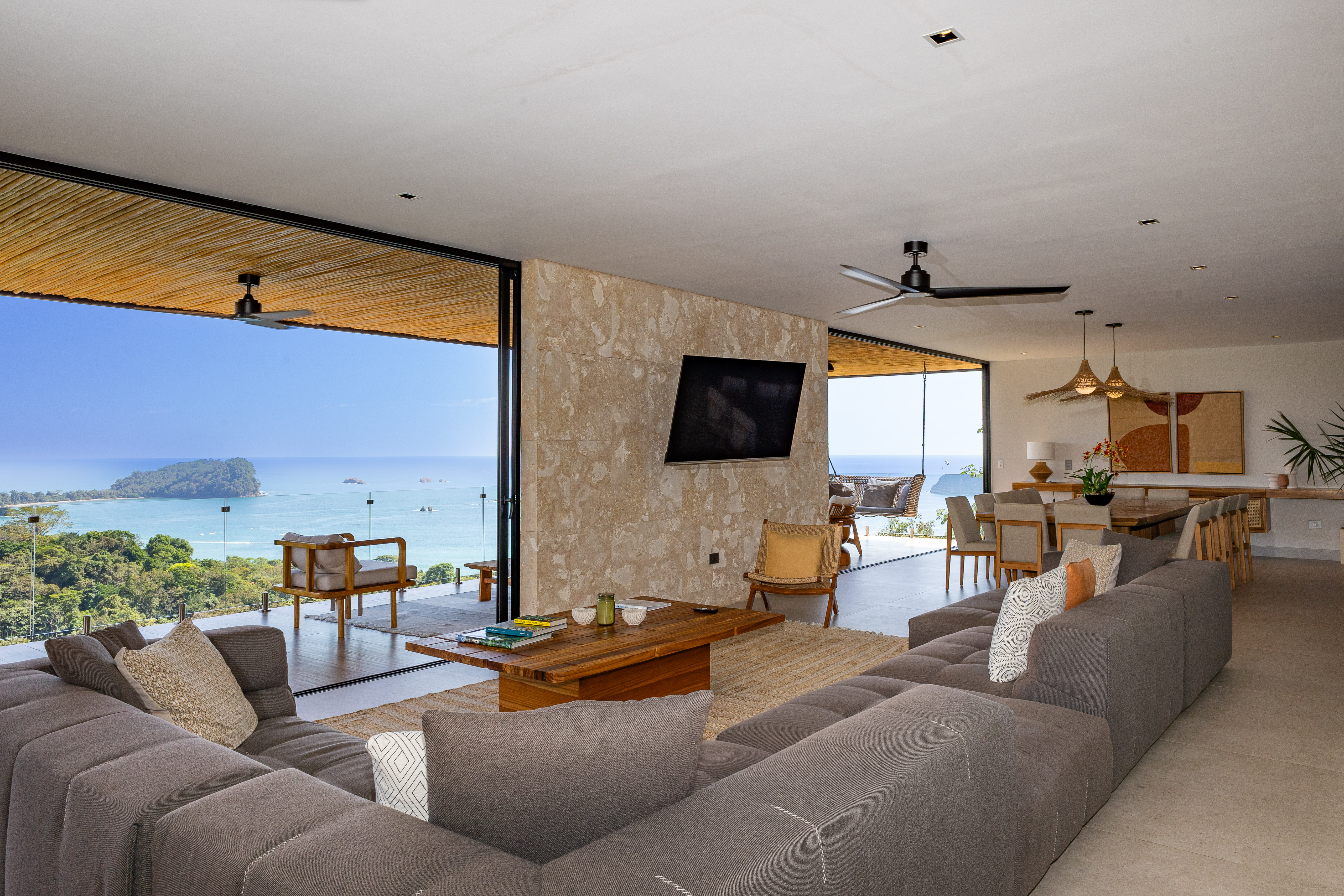 A bright, open-plan living area with floor-to-ceiling glass doors offers panoramic ocean and jungle views, seamlessly connecting the indoor space to an outdoor terrace.