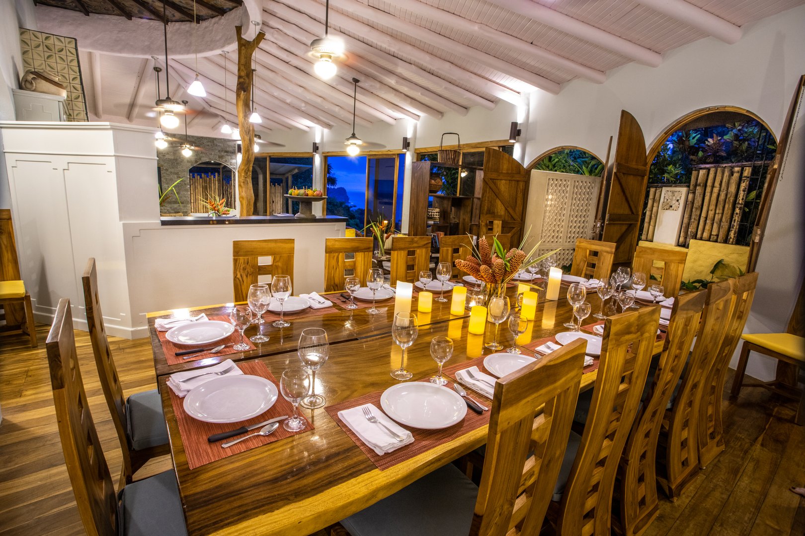The dining room features a long natural wood table, a vaulted ceiling with exposed beams and a tree trunk pillar, and expansive windows looking out to the evening jungle.