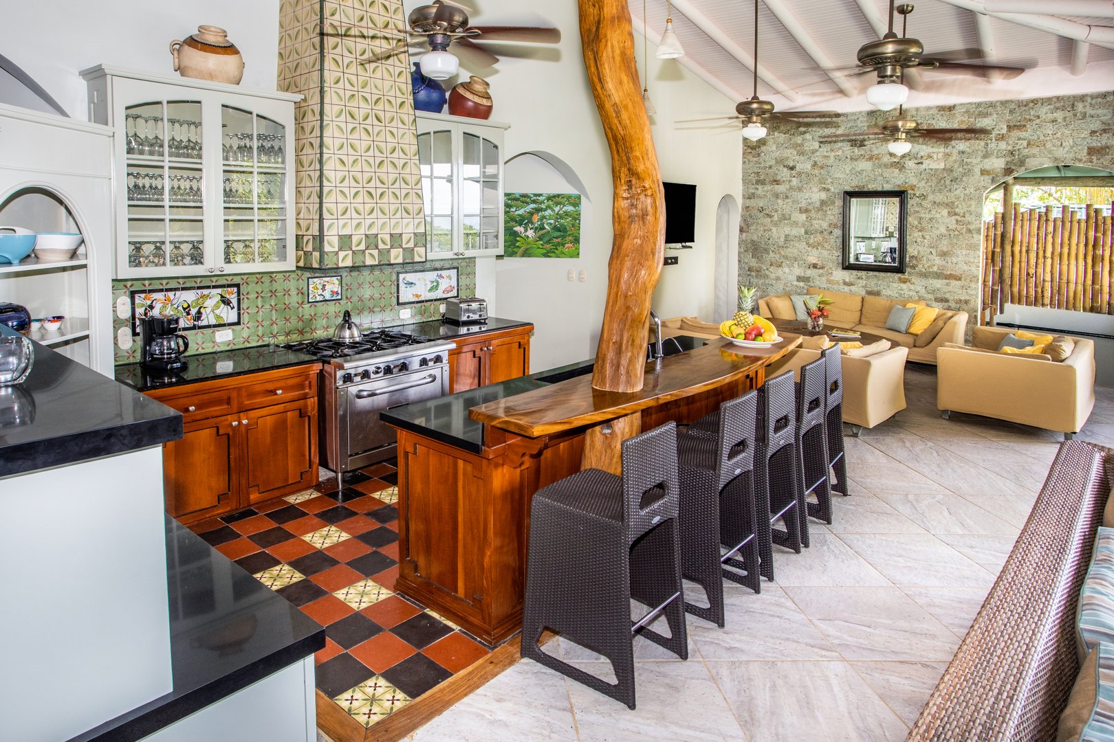A spacious open-concept kitchen with custom tilework and a natural wood bar connects to a living area featuring a stone accent wall, vaulted ceilings, and an archway leading to an outdoor patio.
