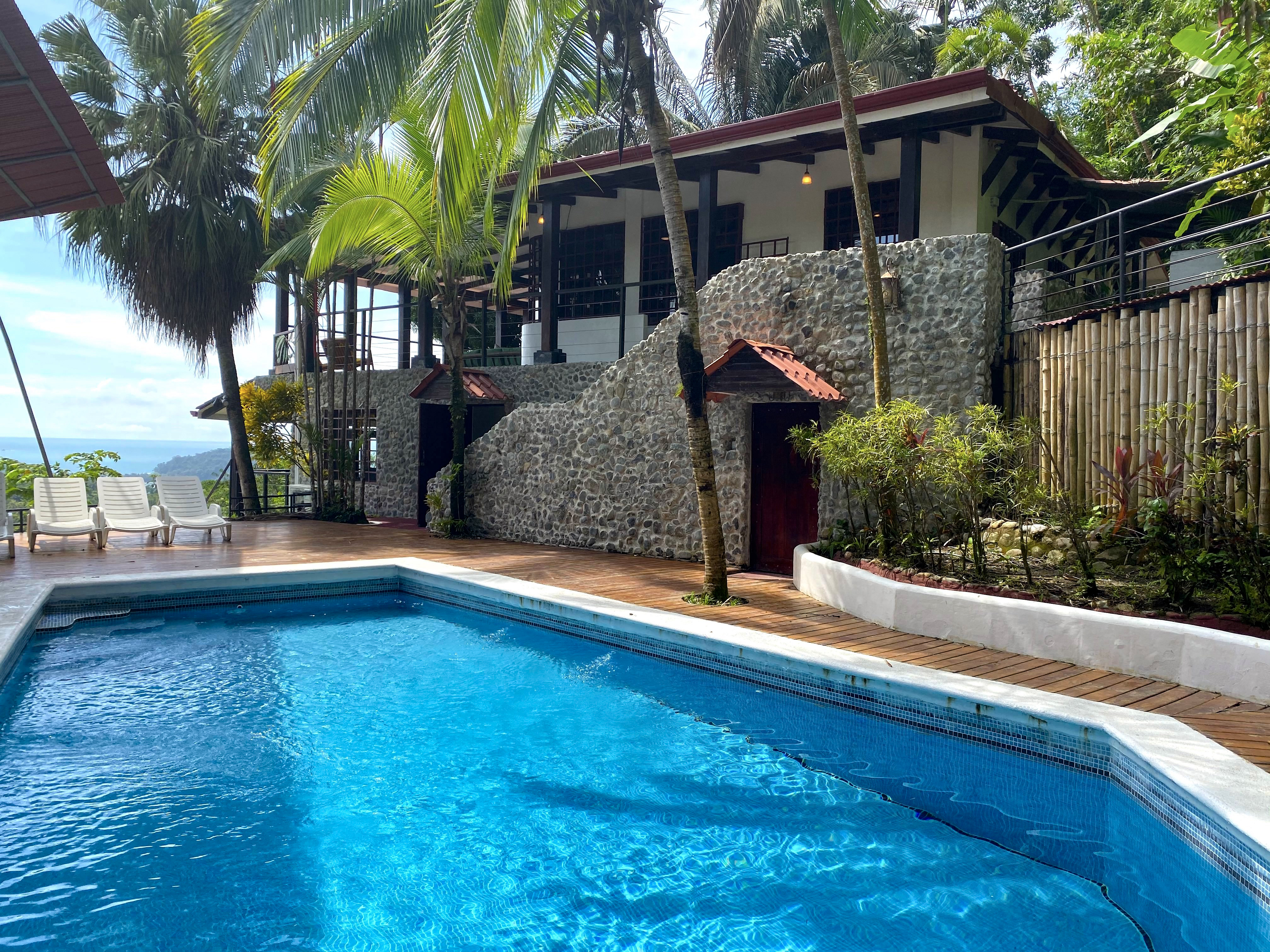 An inviting blue swimming pool with a surrounding wooden deck and lounge chairs overlooks tropical greenery, the villa's stone and wood architecture, and a distant ocean view under bright daylight.