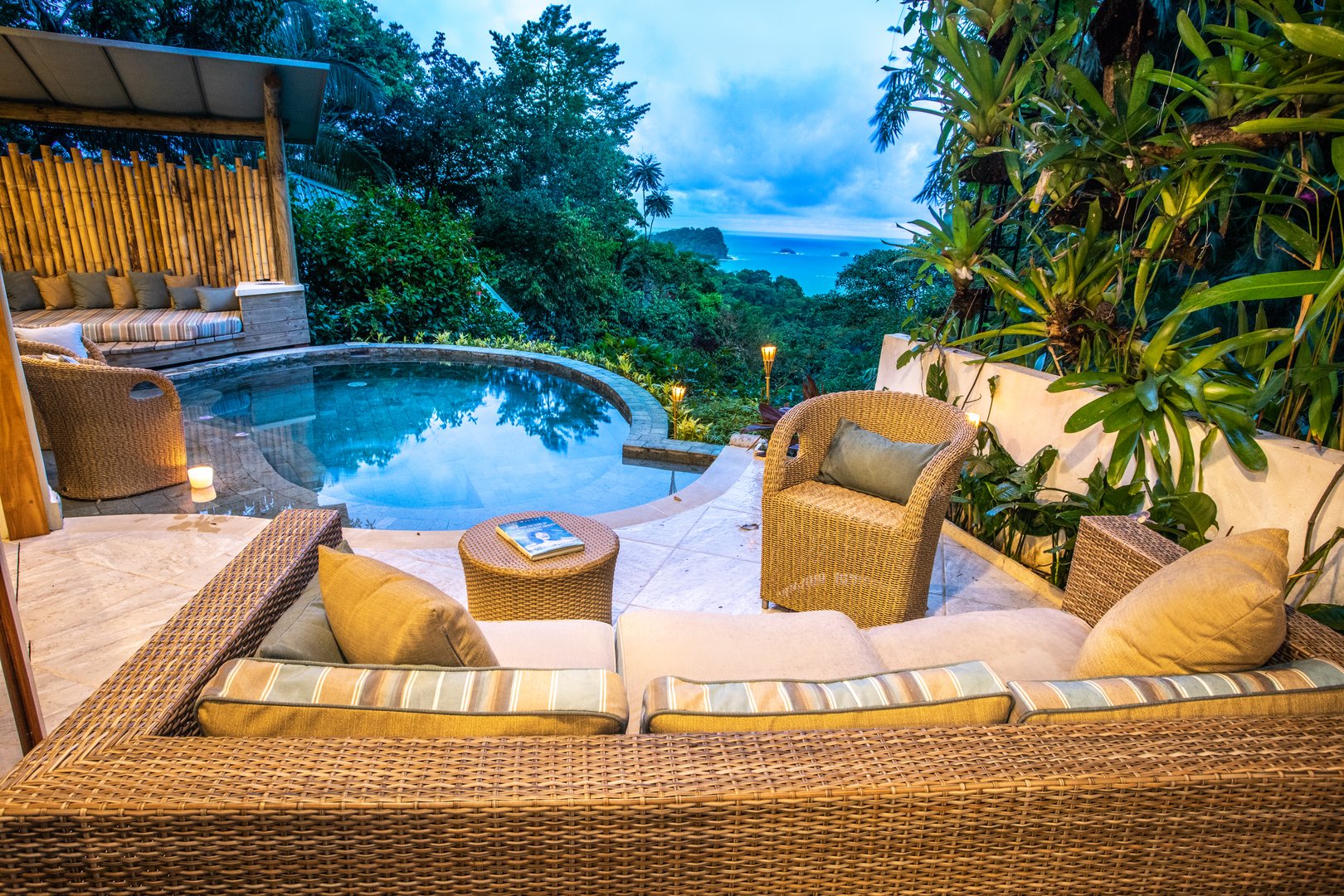 This outdoor terrace offers a circular plunge pool, ample wicker seating, and a bamboo-paneled lounge, all set against a backdrop of lush jungle and distant ocean at dusk.