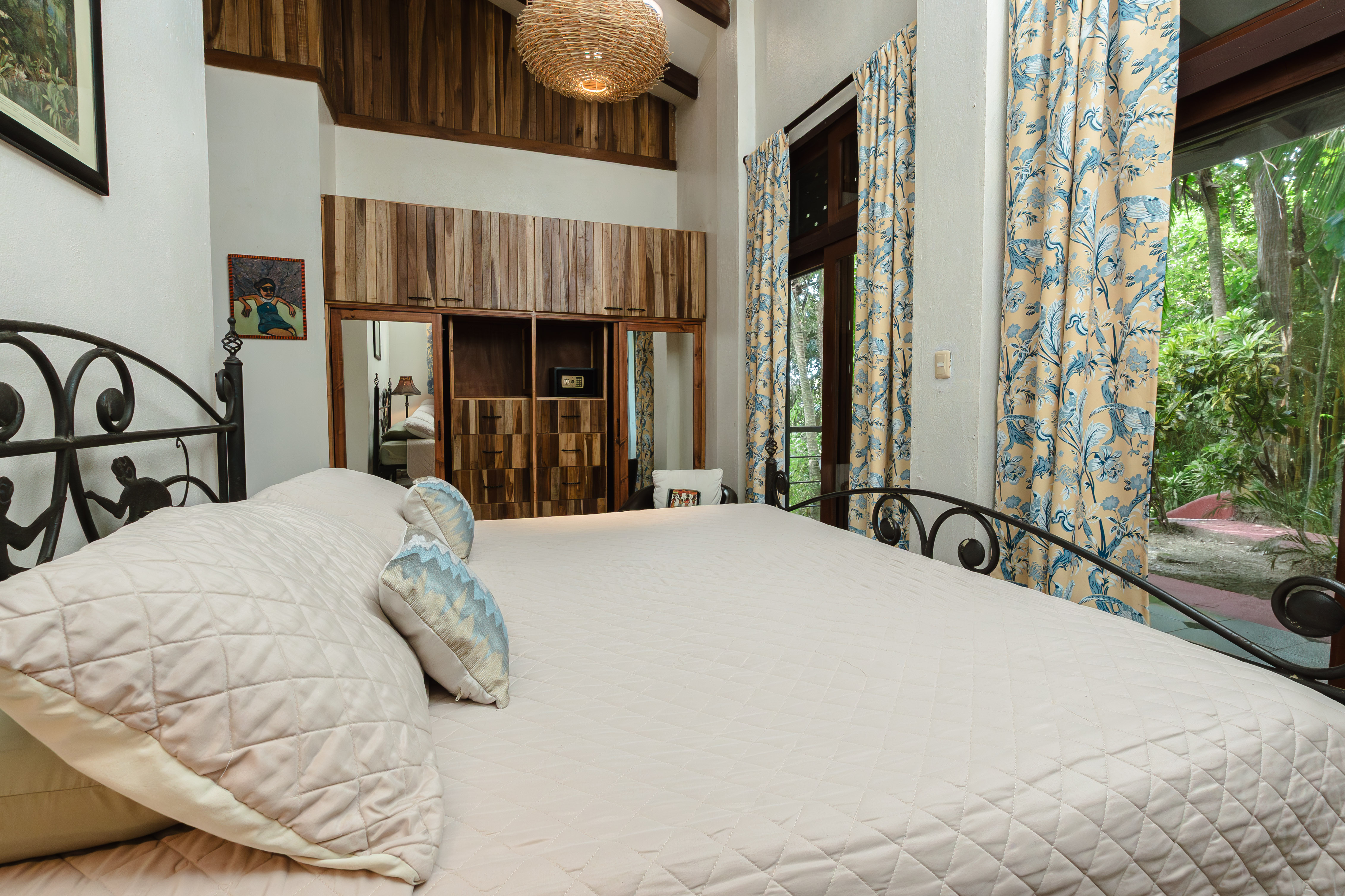 This bright bedroom features a high beamed ceiling with a woven light fixture, a large metal-framed bed, built-in wooden storage, and a glass door looking out to tropical greenery.