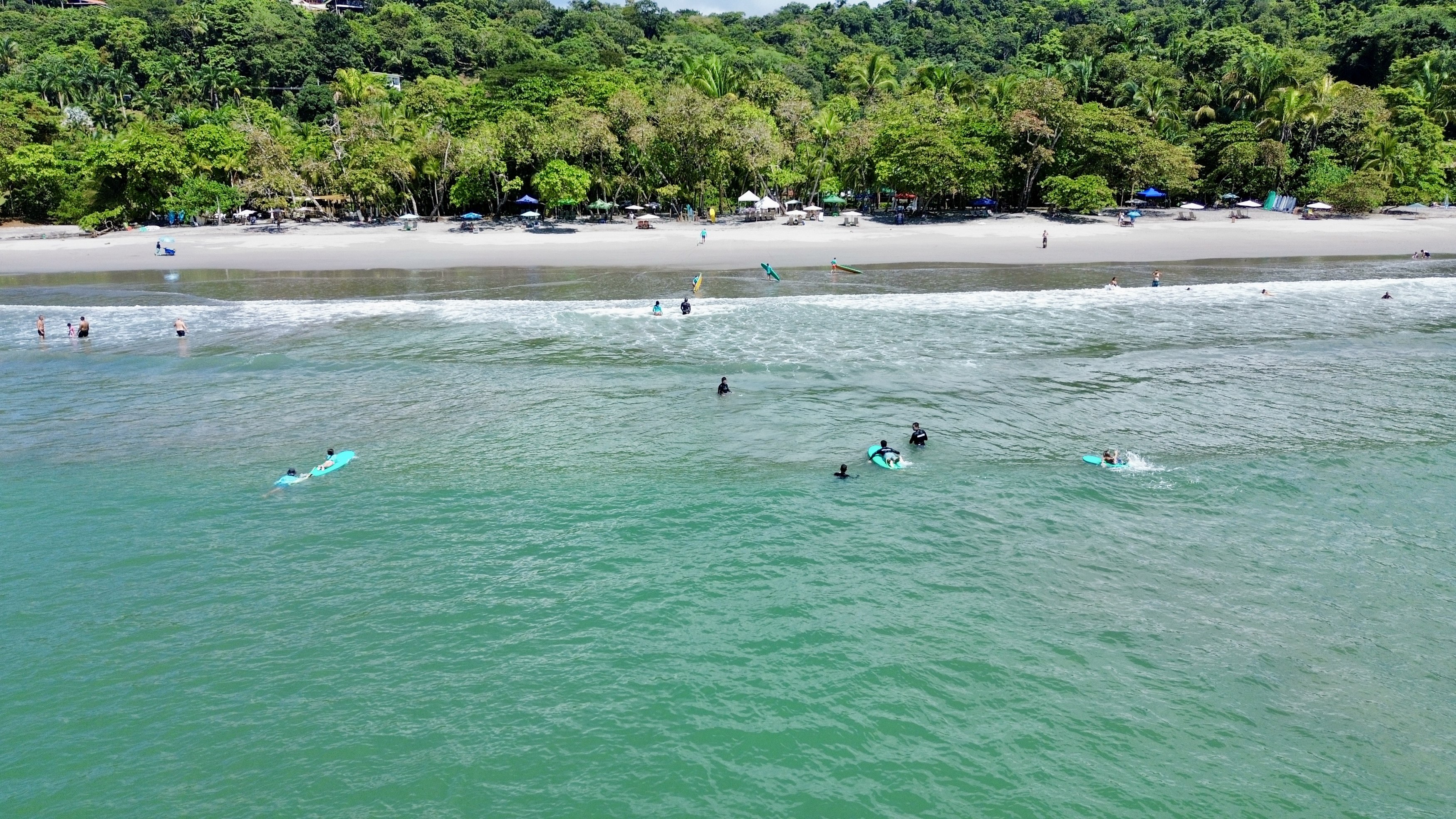 An aerial view showcases the turquoise Pacific Ocean with people swimming and surfing, a sandy beach with umbrellas, and a dense, verdant jungle-covered hillside.