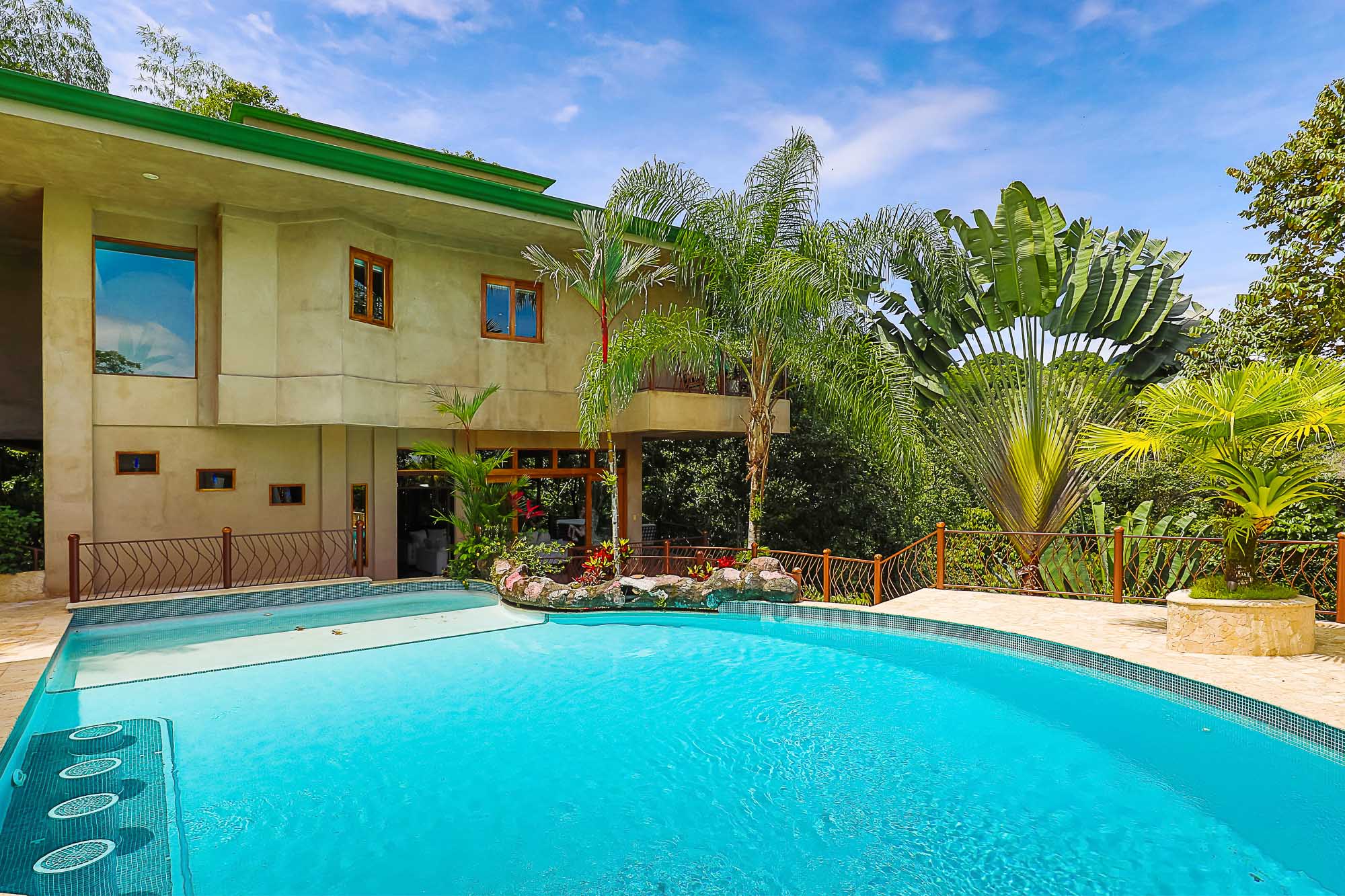 The multi-level villa's facade overlooks an extensive outdoor swimming pool and sun deck, all enveloped by tropical rainforest under a bright sky.