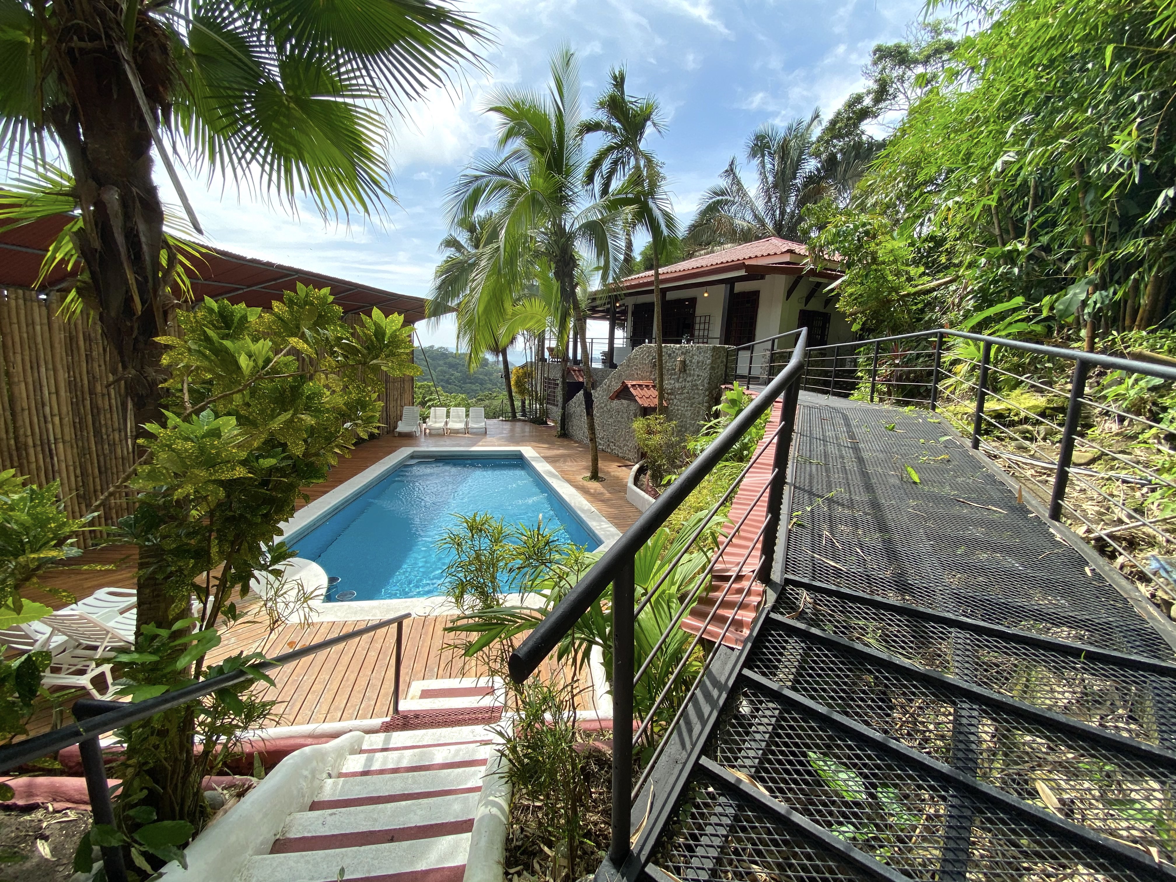 The inviting blue swimming pool with a surrounding wooden deck and sun loungers is set amidst lush tropical foliage, offering views of green hills and access to the villa through multiple pathways.