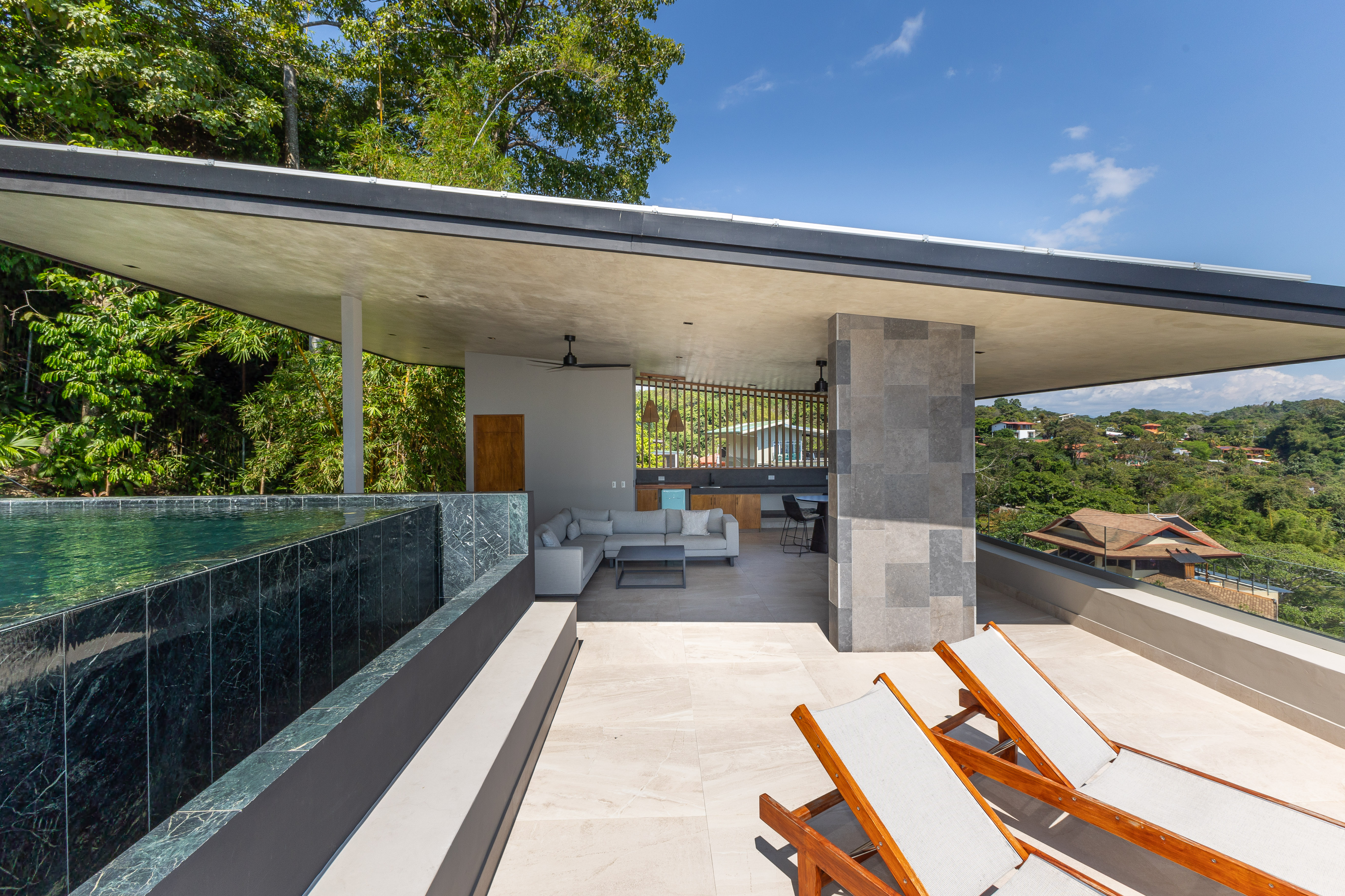 A rooftop infinity pool and a covered outdoor living area with kitchen are surrounded by tropical jungle and distant hillside views under a bright sky.