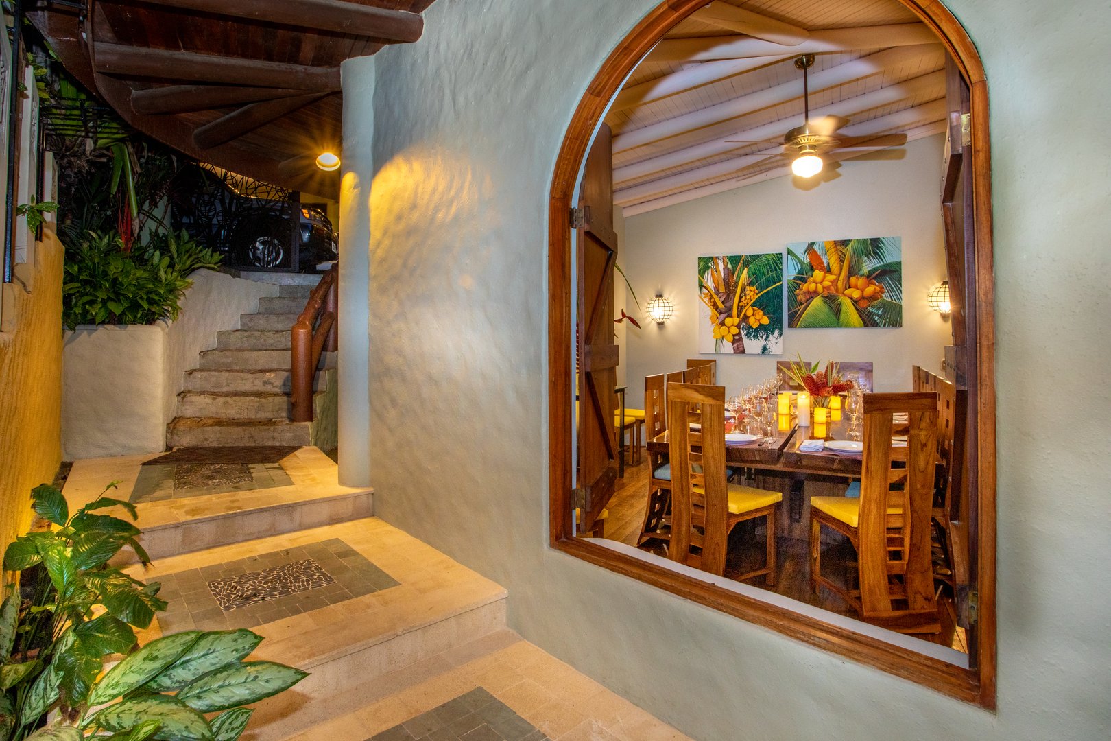 An arched wooden entryway frames an inviting dining room featuring a high wood-paneled ceiling, tropical artwork, and a long table, situated next to an illuminated outdoor staircase.