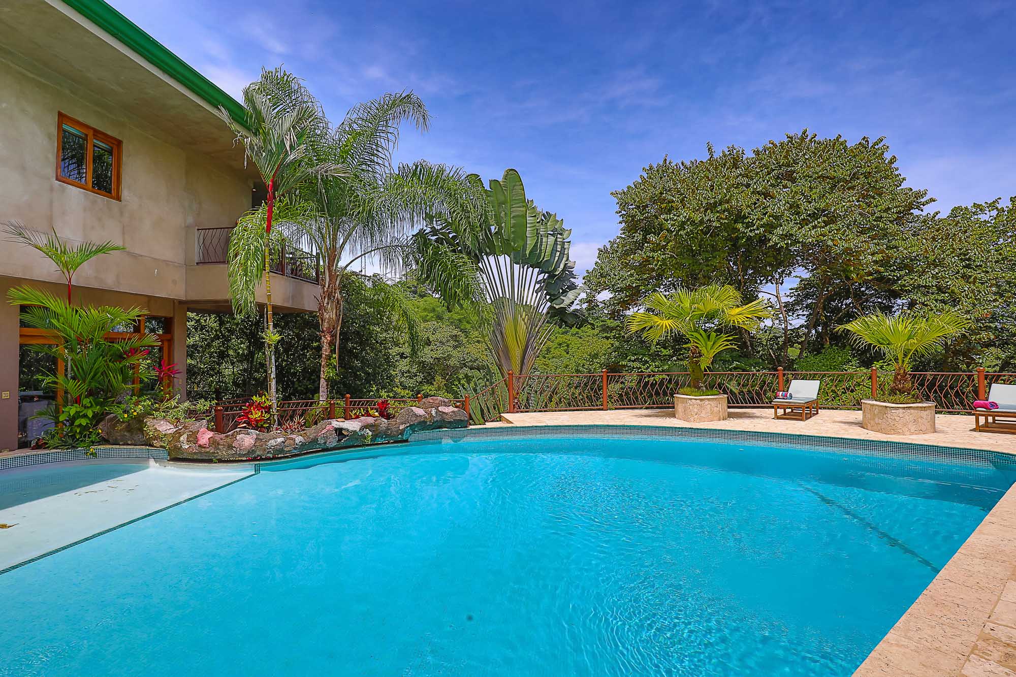 A bright blue outdoor swimming pool is set against a multi-level villa with wood-framed windows and surrounded by lush tropical rainforest under a sunny sky.