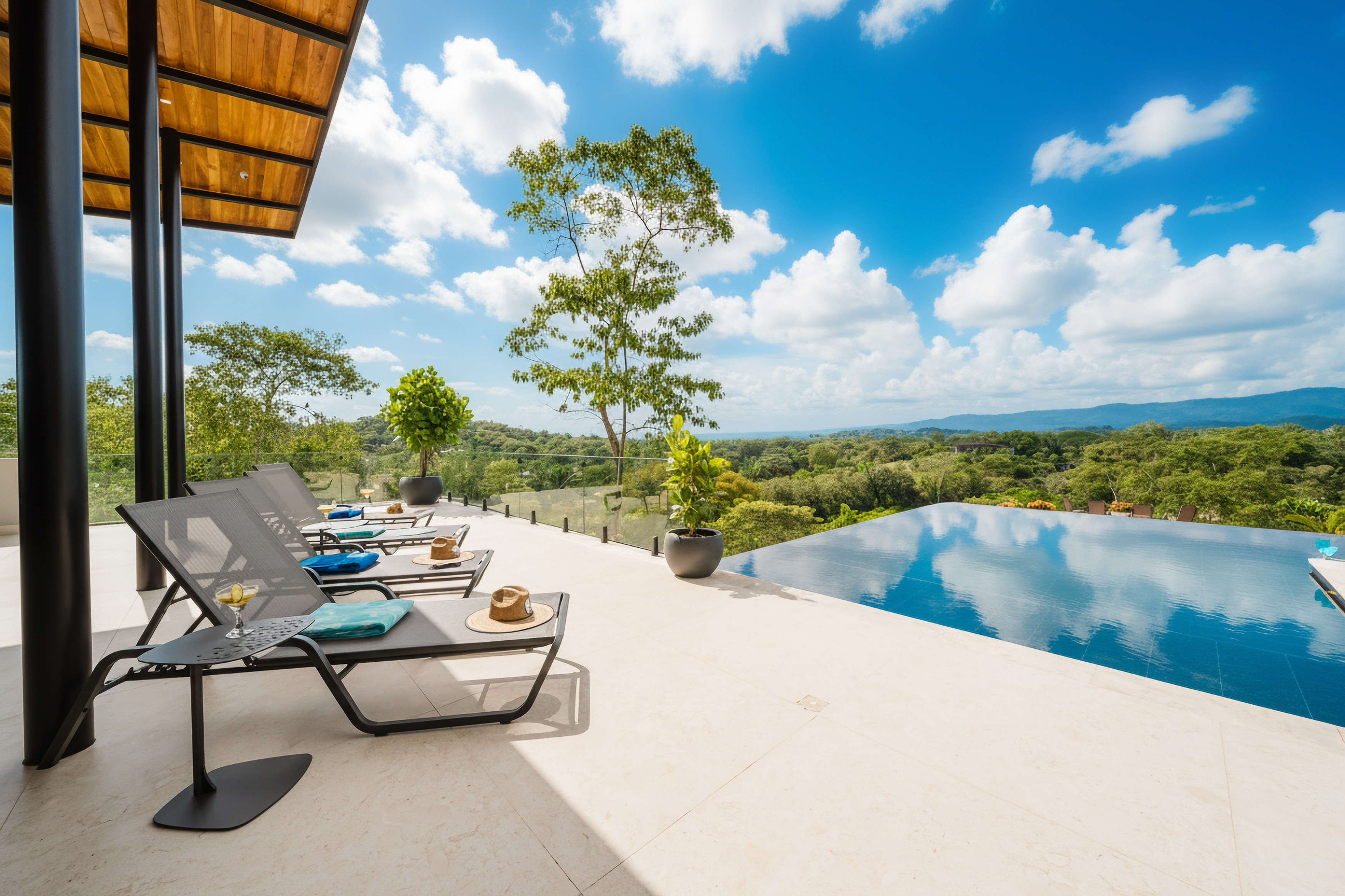 A tiled terrace with shaded lounge chairs extends to an infinity pool, overlooking a dense green landscape under a bright sky.