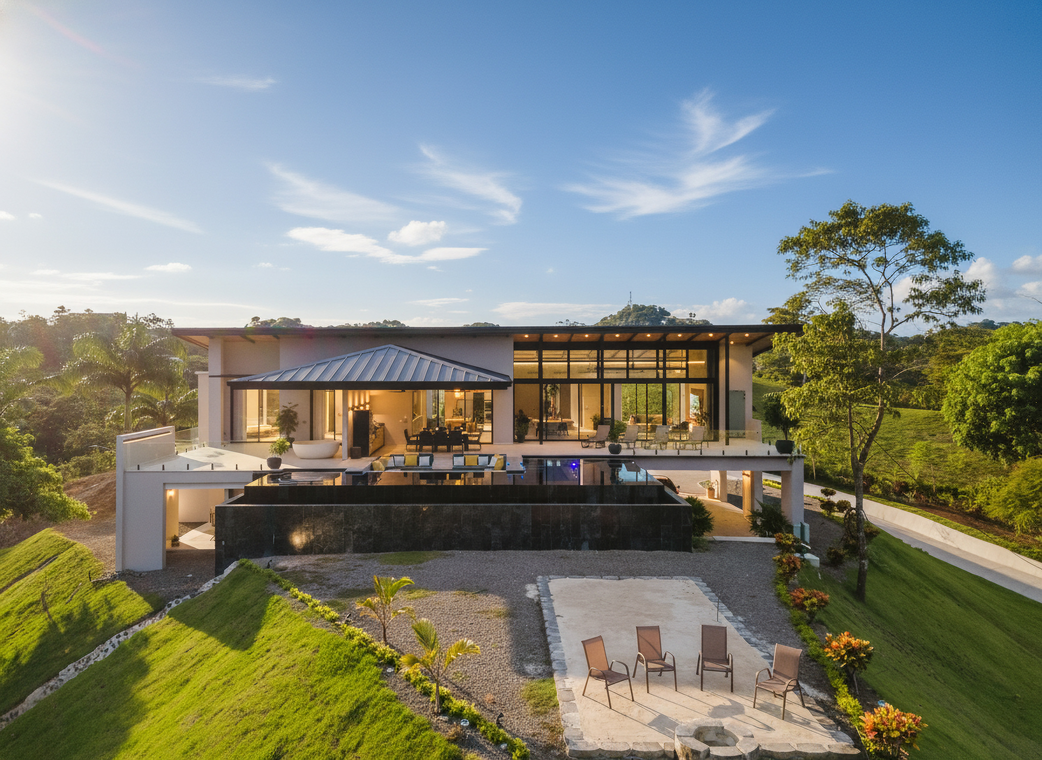 This modern multi-level villa features expansive glass walls and an infinity pool that overlooks a lush tropical forest and distant hills under a soft daylight sky.