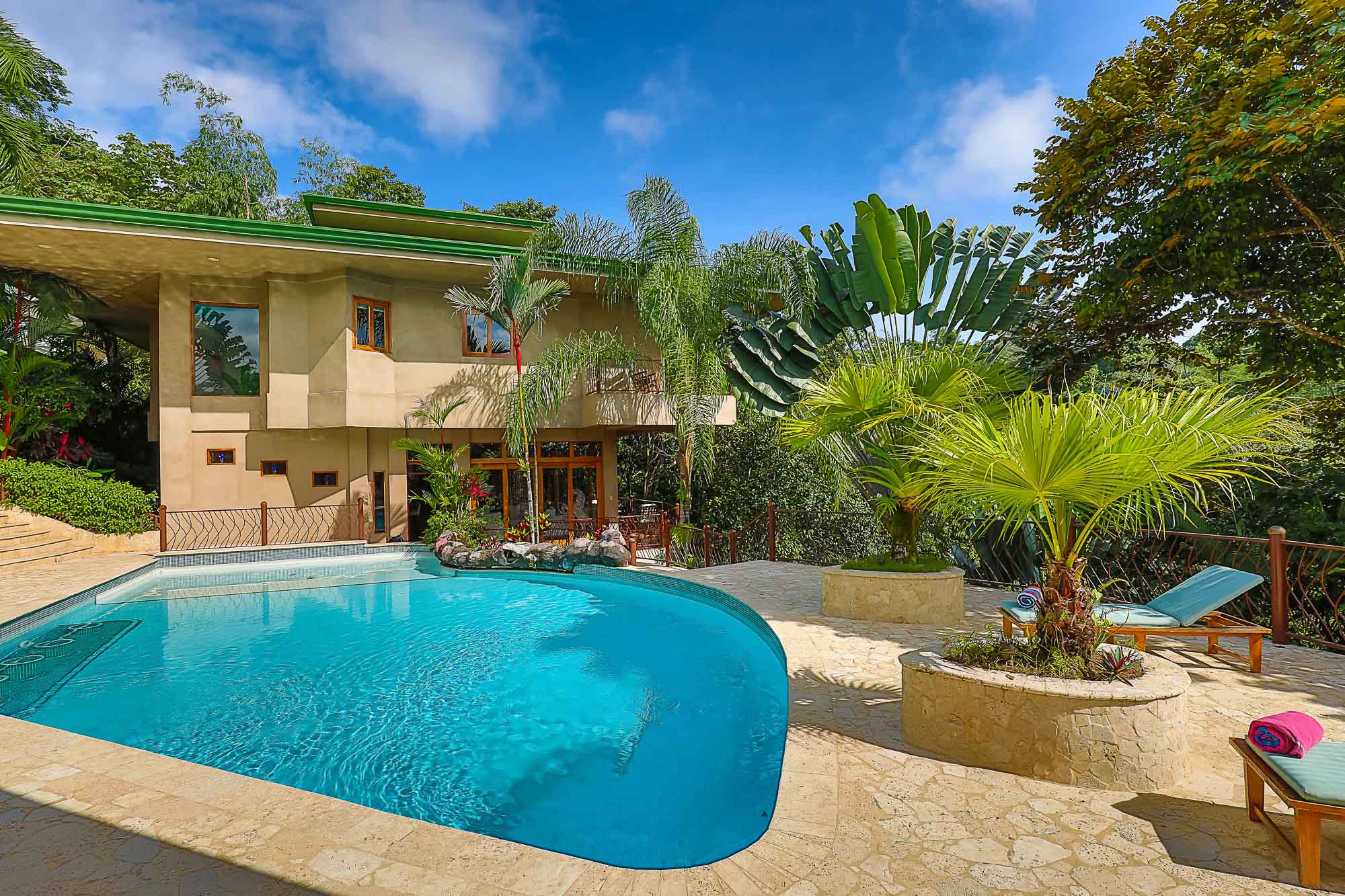 A large, curved swimming pool and stone patio are visible in front of a multi-level modern villa, surrounded by dense tropical foliage under a bright blue sky.