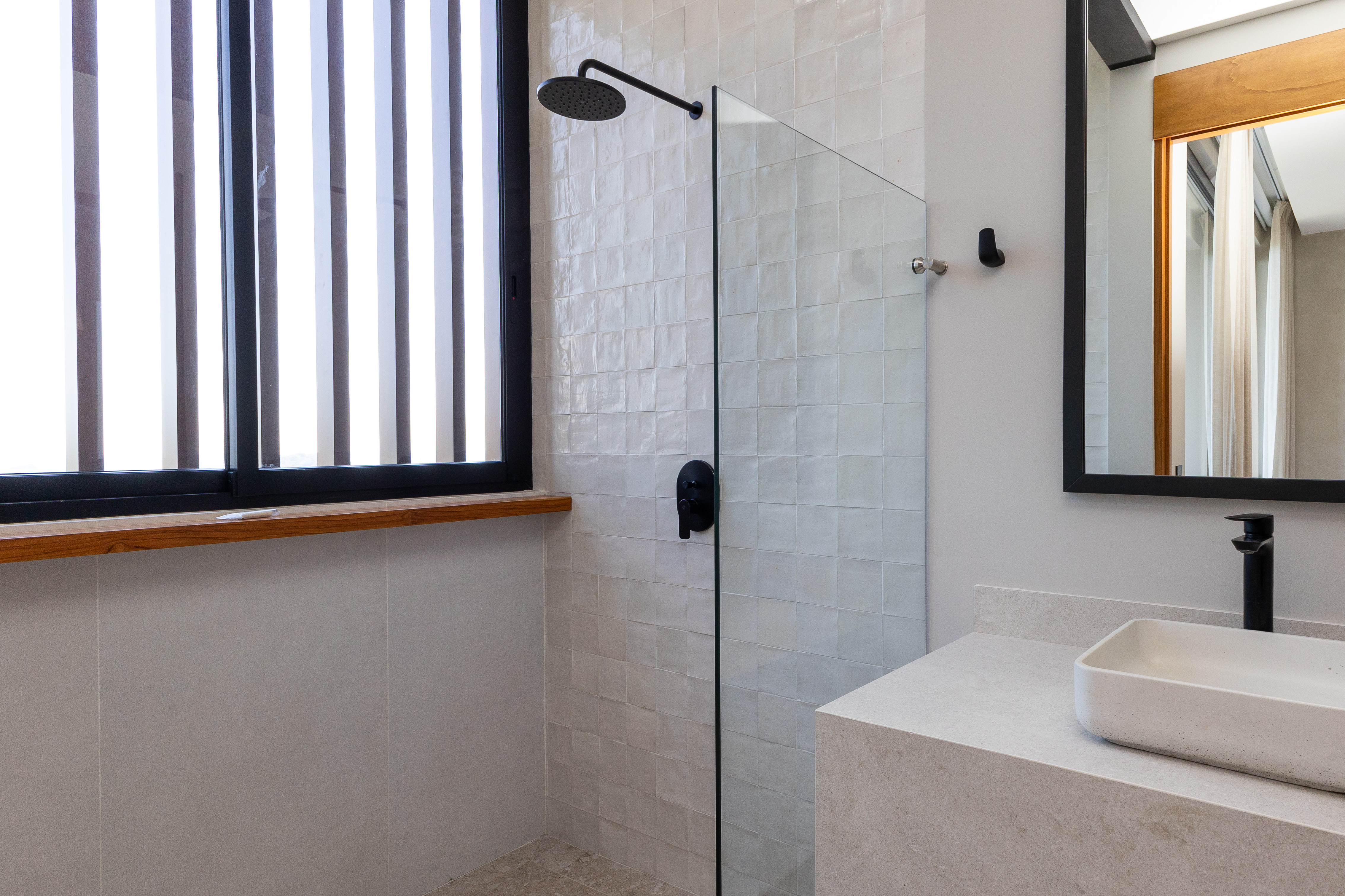 The bathroom features a walk-in shower with matte black fixtures and textured white tiles, a stone vanity with a rectangular sink, and natural light from a louvered window.