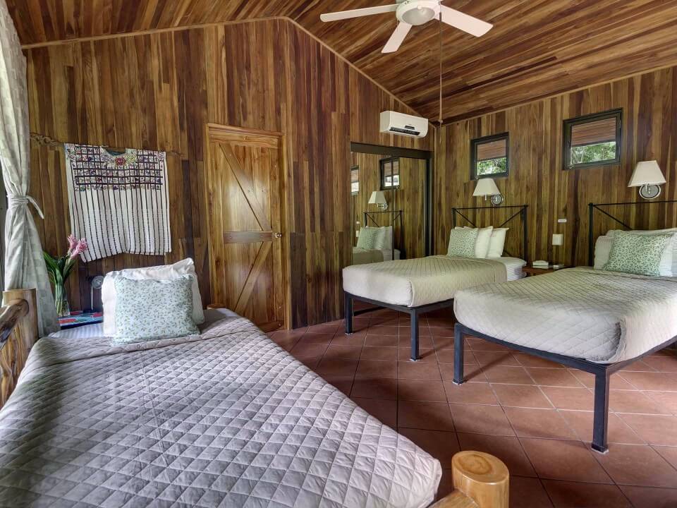 A bedroom with warm wood-paneled walls and a vaulted ceiling features three beds, terracotta-tiled floors, and windows looking out to jungle greenery.