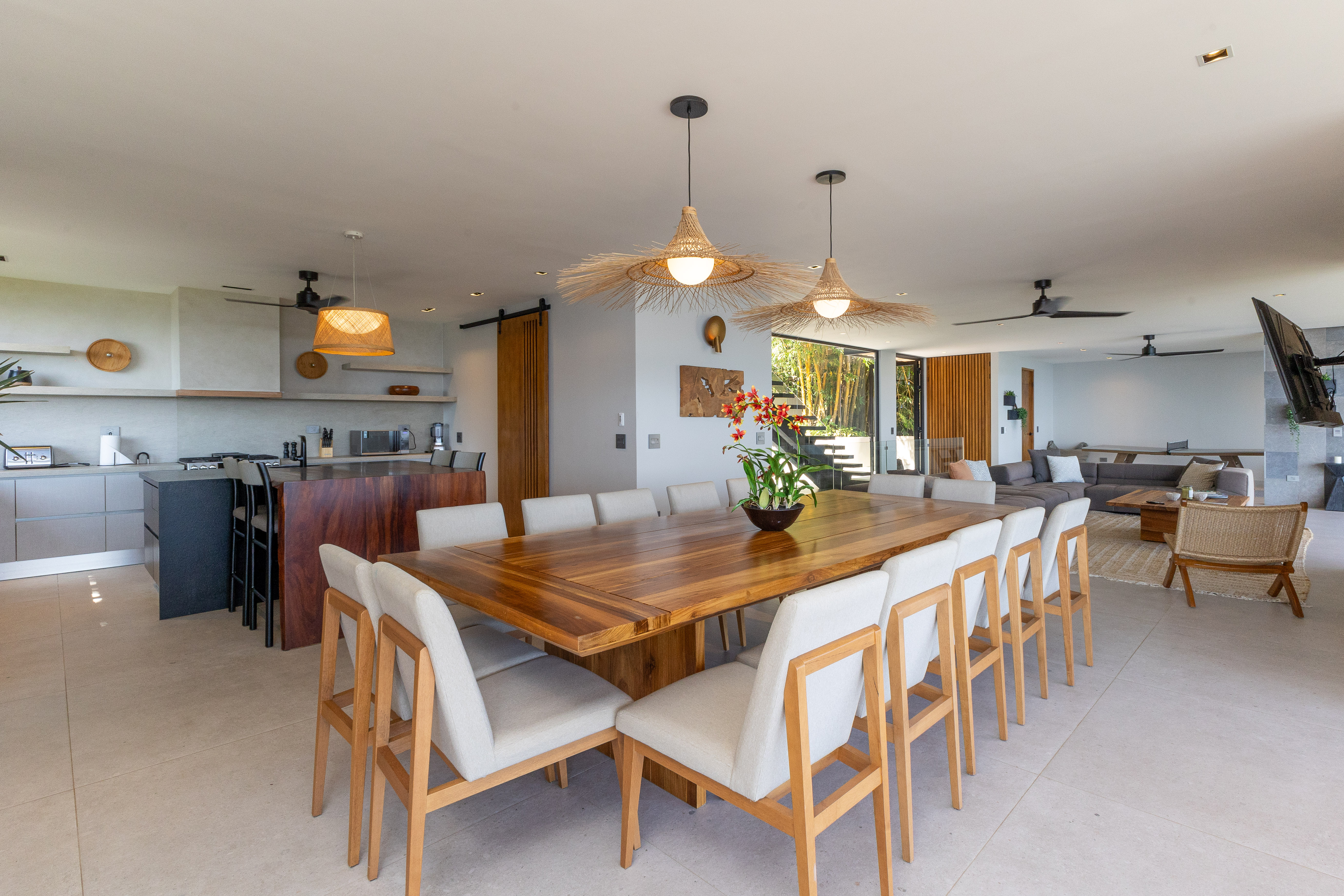 A spacious open-plan dining area with a large wooden table and chairs flows into a modern kitchen and lounge, offering natural light and views of the surrounding jungle through large glass doors.