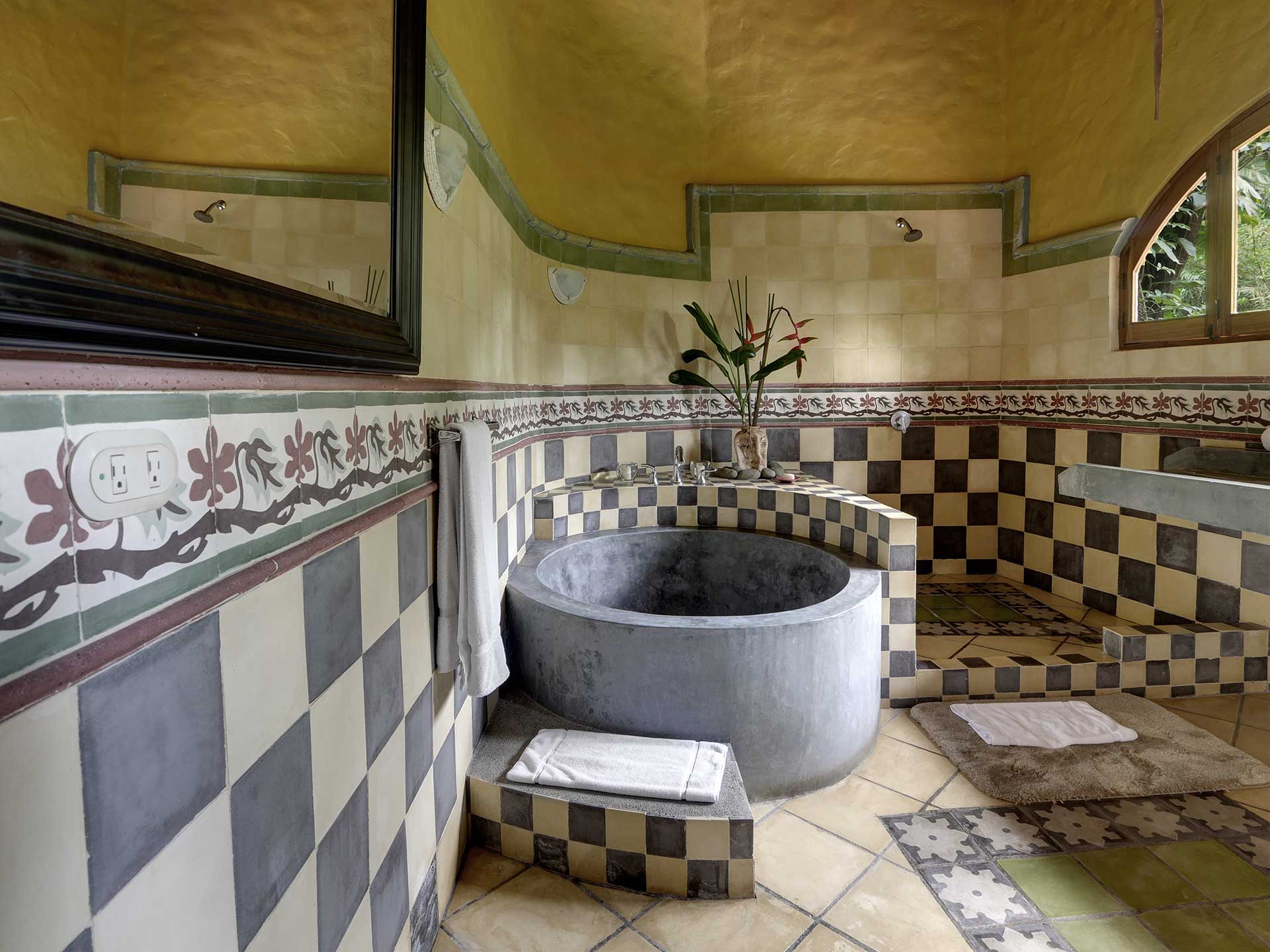 A custom-tiled bathroom with a deep round soaking tub and walk-in shower features an arched window overlooking lush jungle.
