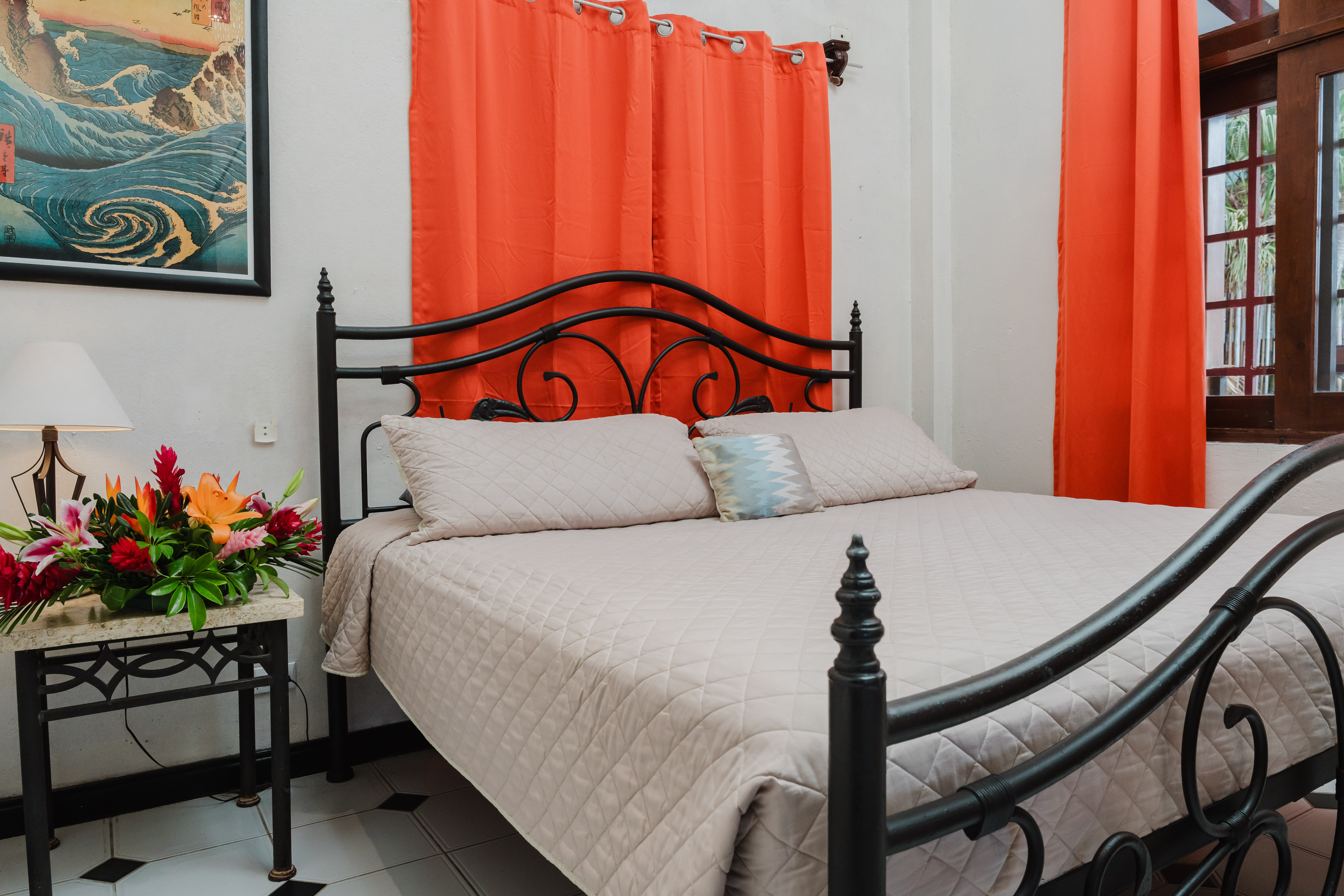 This bedroom features a wrought iron bed, with bright orange curtains drawn over a window that offers filtered natural light and a view of tropical foliage.