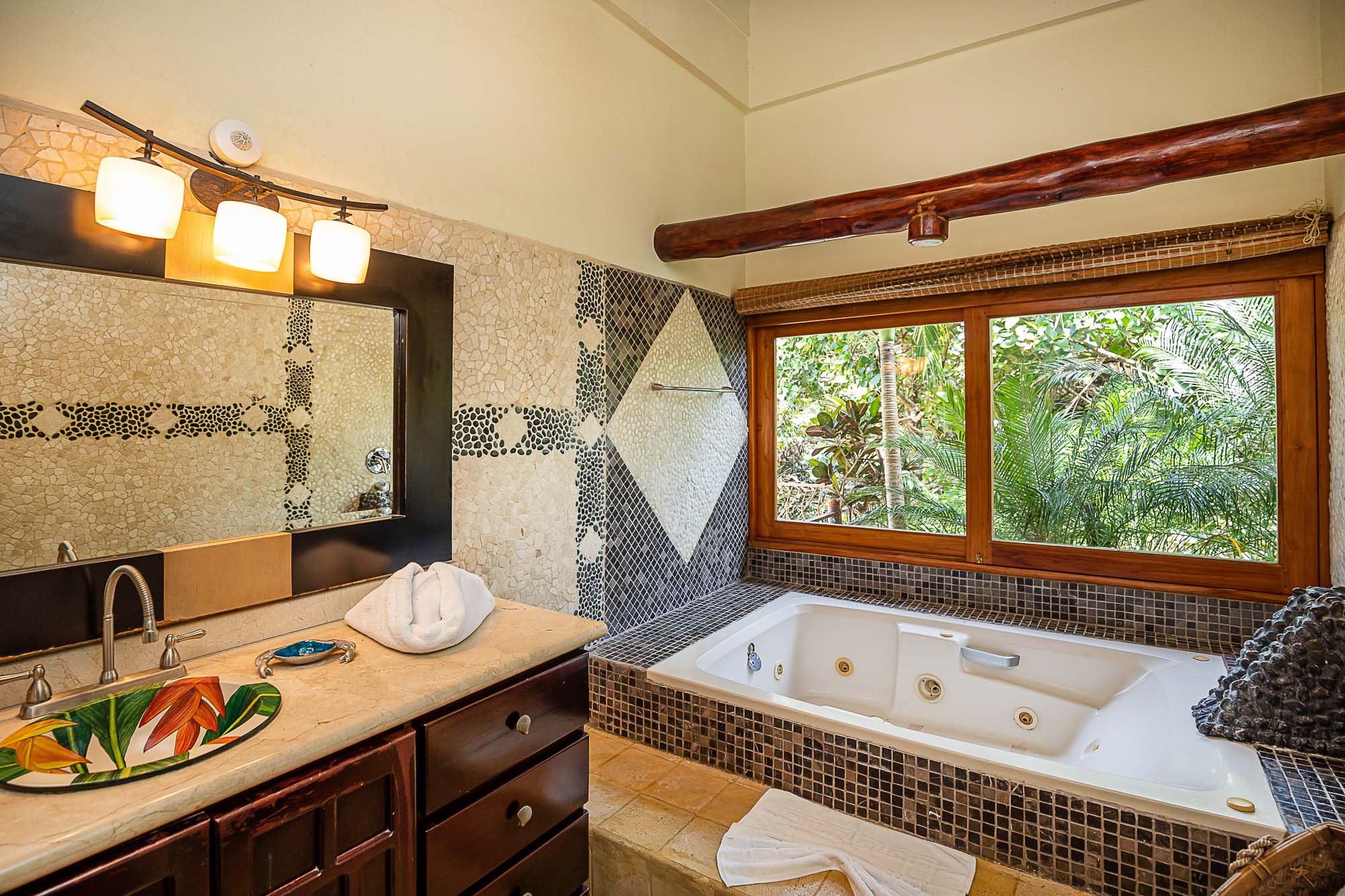 A bathroom features a jetted tub positioned by a large window offering natural light and views of lush tropical foliage, complemented by a rustic wooden beam and decorative mosaic tile walls.
