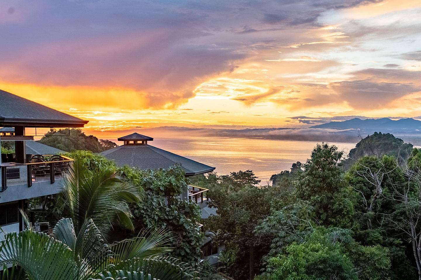 A vibrant sunset over the Pacific Ocean is visible from the villa's balconies, surrounded by lush tropical rainforest.
