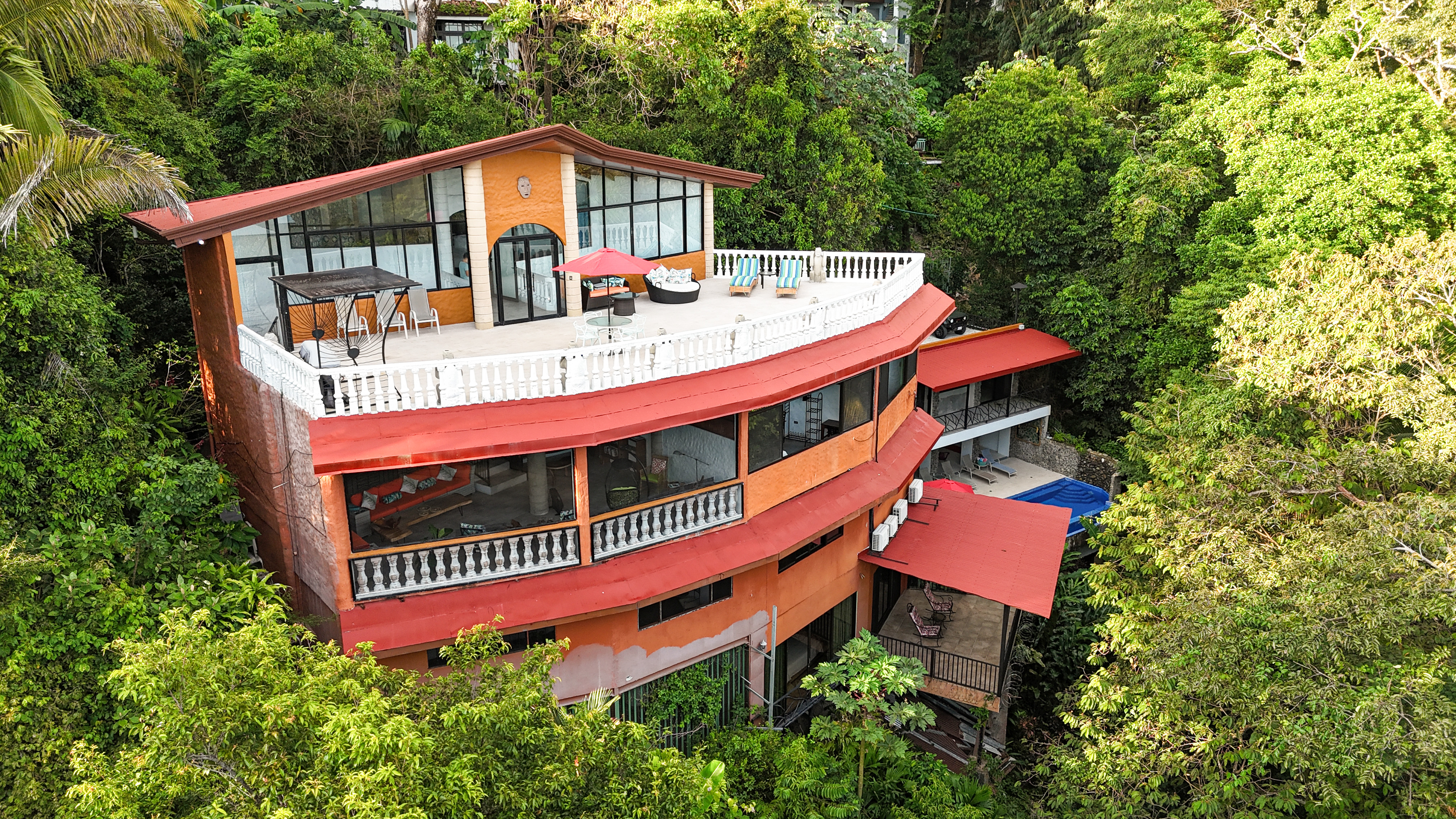 An aerial view shows a multi-level villa with orange walls, red roofs, and several terraces with white balustrades, deeply set within a tropical jungle, with an outdoor pool visible on a lower level.