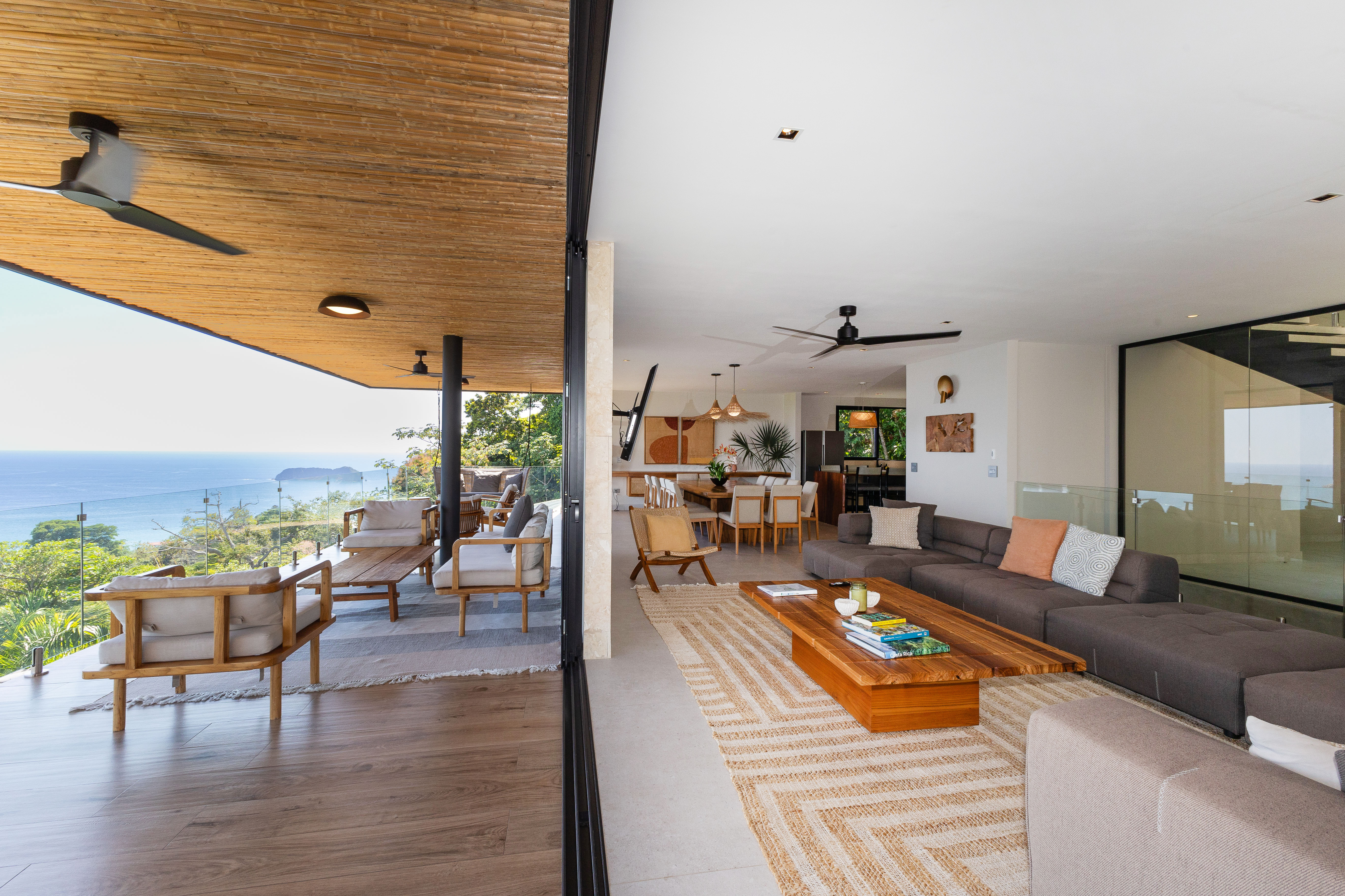 An open-concept living area with a large sofa flows directly to a furnished outdoor terrace through wide glass doors, framing views of the ocean and surrounding jungle.