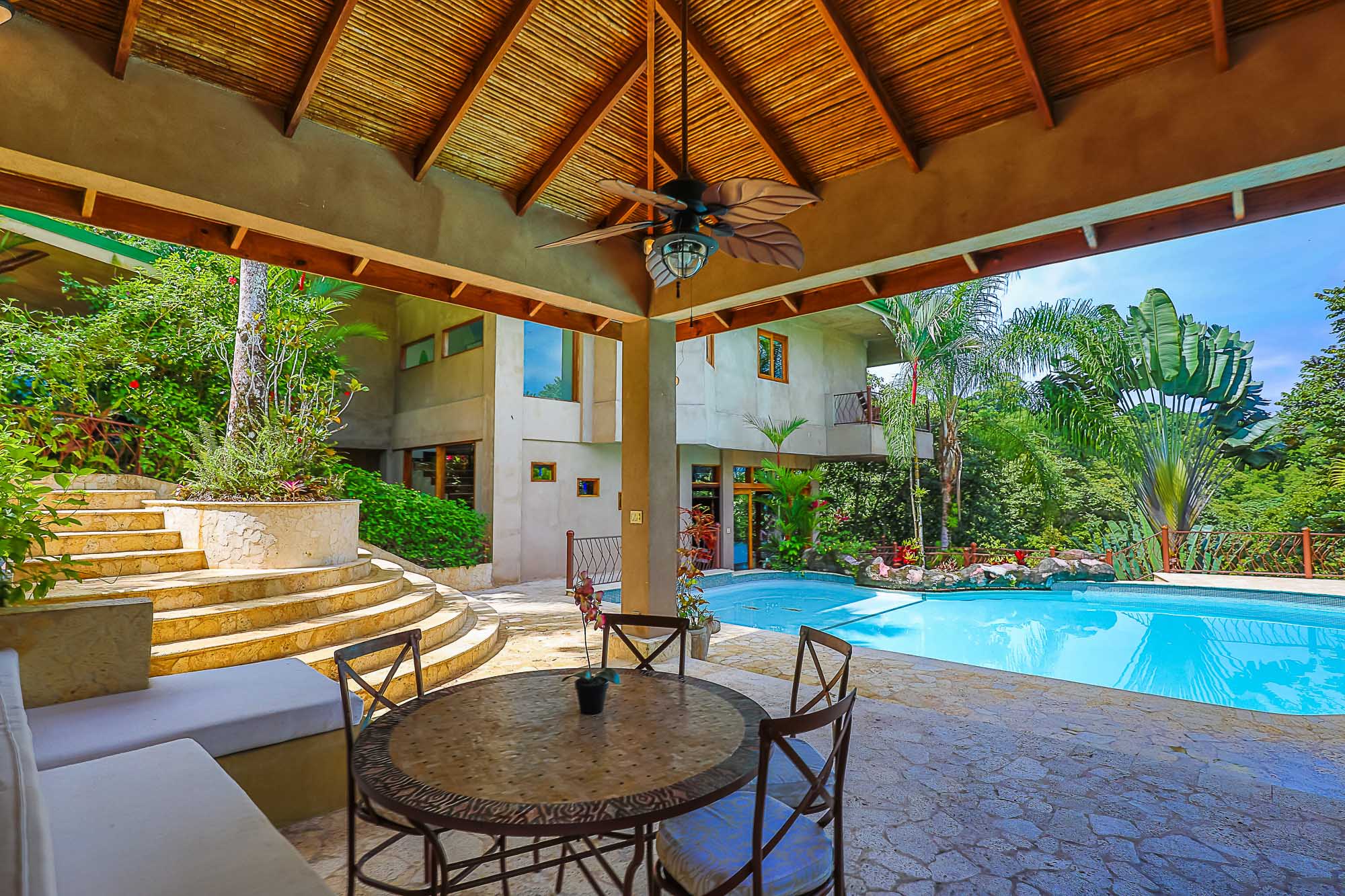 A shaded poolside terrace features a high slatted wood ceiling and an open dining area, offering direct access to the pool and views of the villa set against lush tropical landscaping.