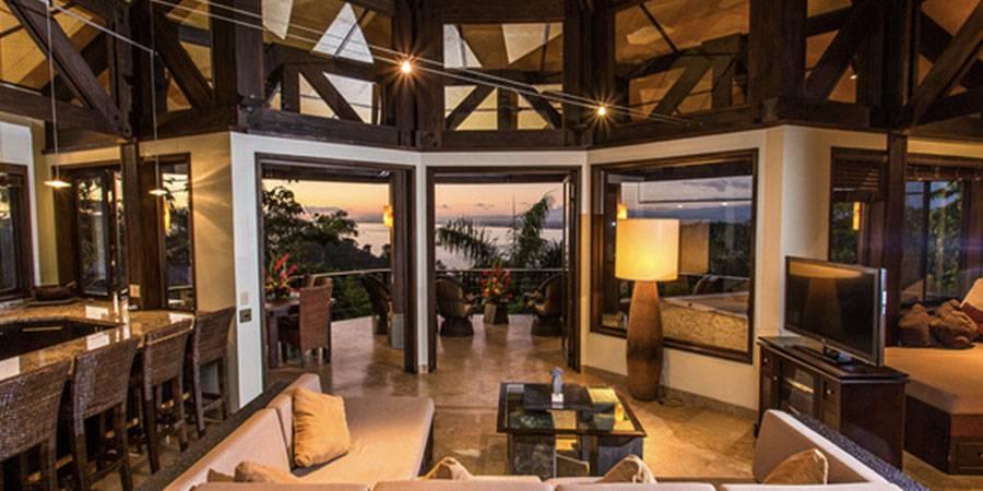 An open-plan living area with high beamed ceilings and an integrated kitchen bar features large glass doors opening to a balcony with an ocean and sunset view.