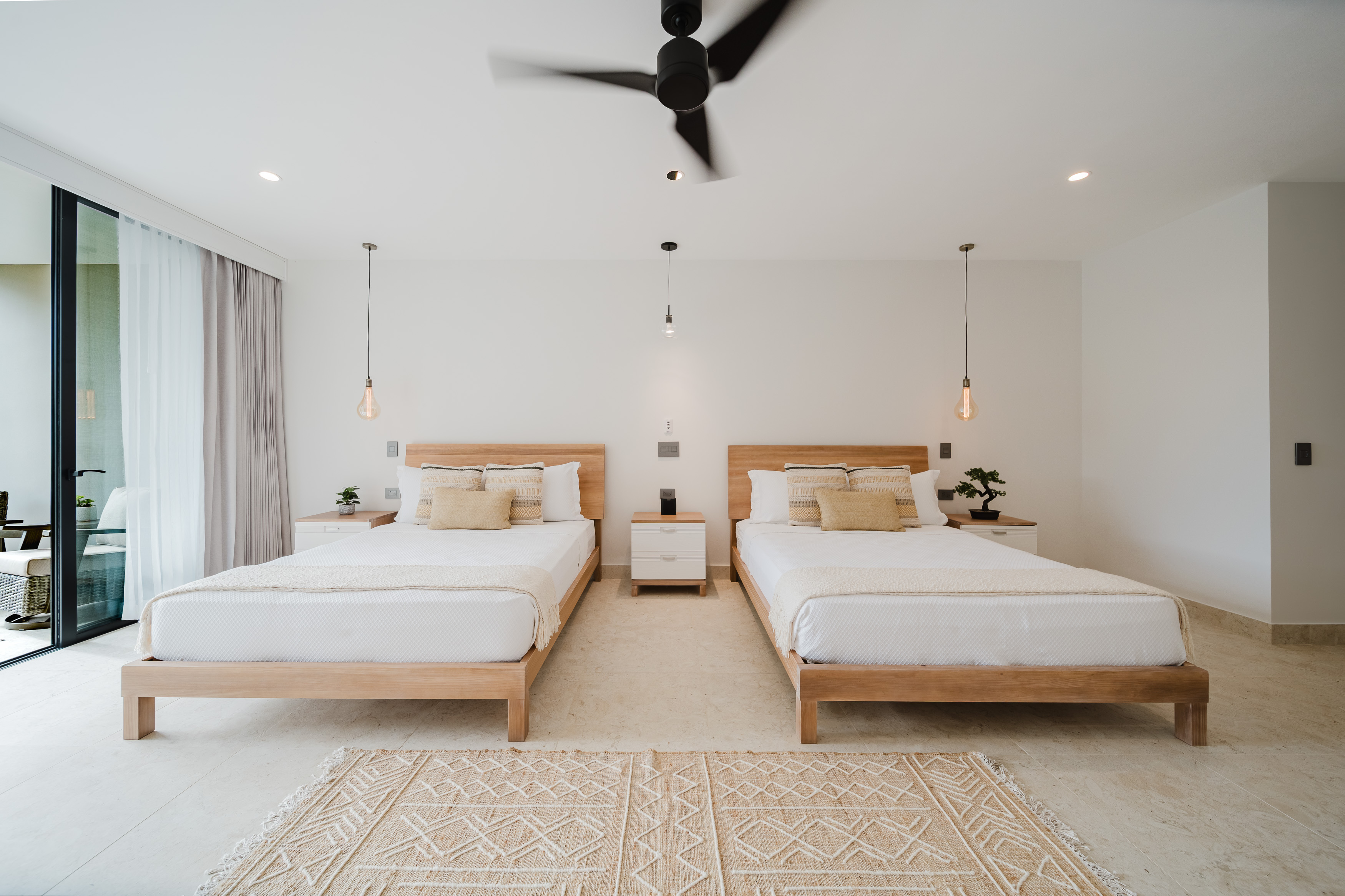 A bright bedroom features two wooden-framed beds, a light tiled floor, and a large sliding glass door opening to an outdoor area, complemented by a ceiling fan and pendant lighting.
