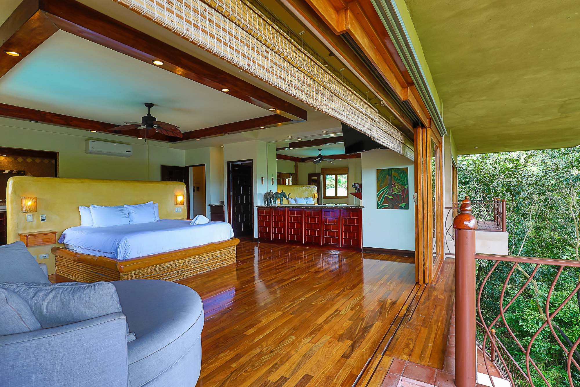 This spacious bedroom features warm wooden floors and ceiling beams, a large bed, and wide folding glass doors that open to a private terrace with direct views of the lush tropical rainforest.
