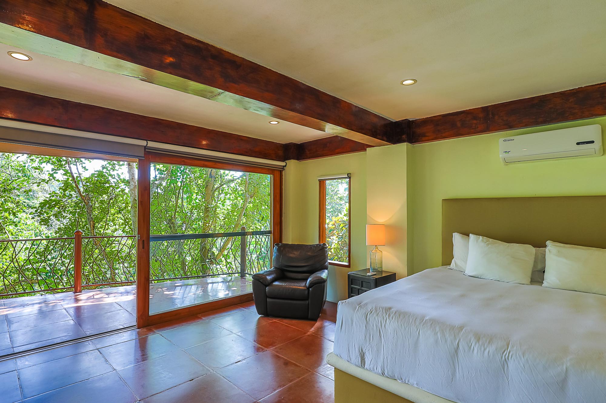 This bedroom offers ample natural light and direct rainforest views through its large sliding glass doors, complemented by exposed wooden ceiling beams and terracotta floor tiles.