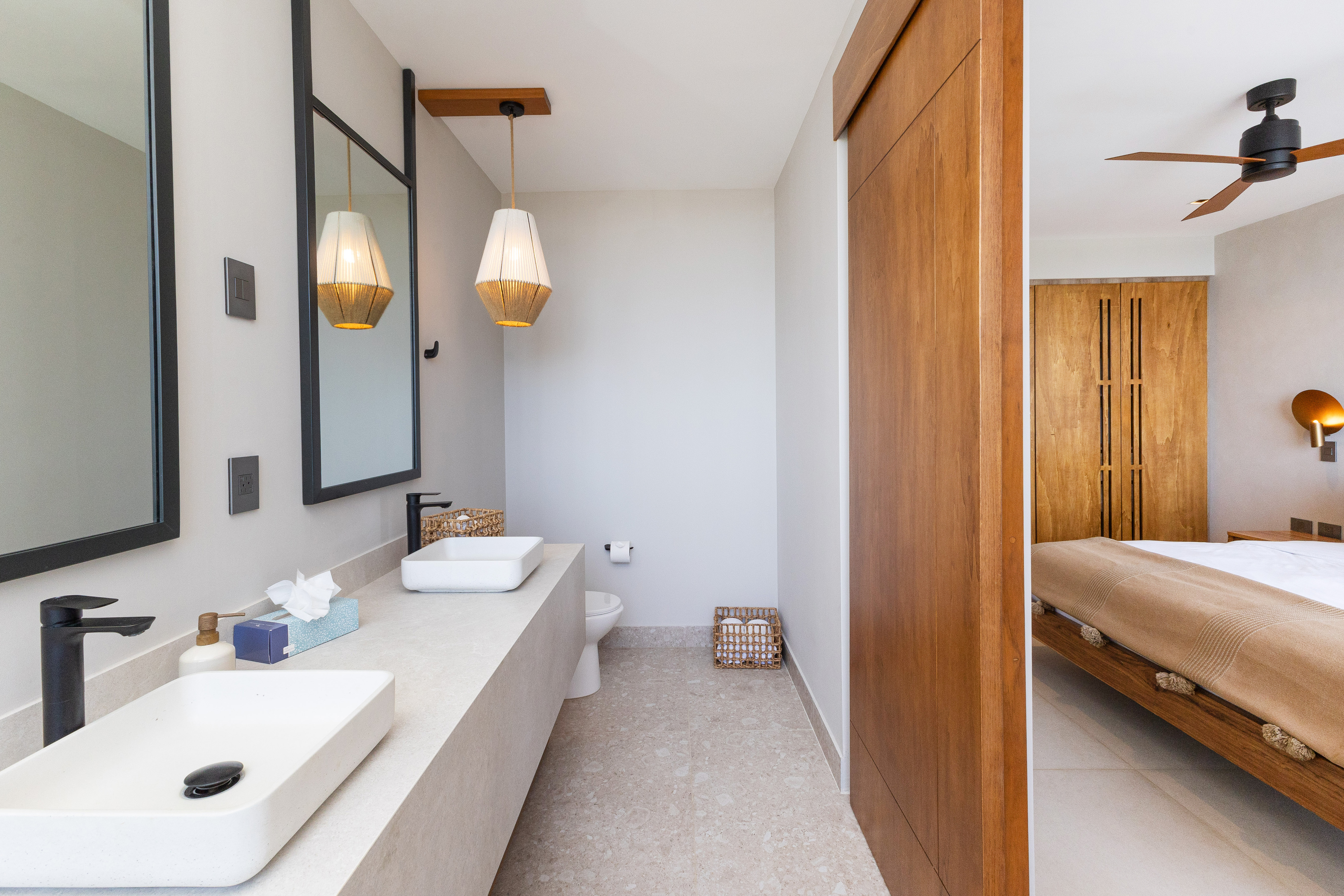 A contemporary bathroom with dual vessel sinks, dark fixtures, and framed mirrors is separated by a natural wood sliding door from a bedroom featuring a bed and ceiling fan.