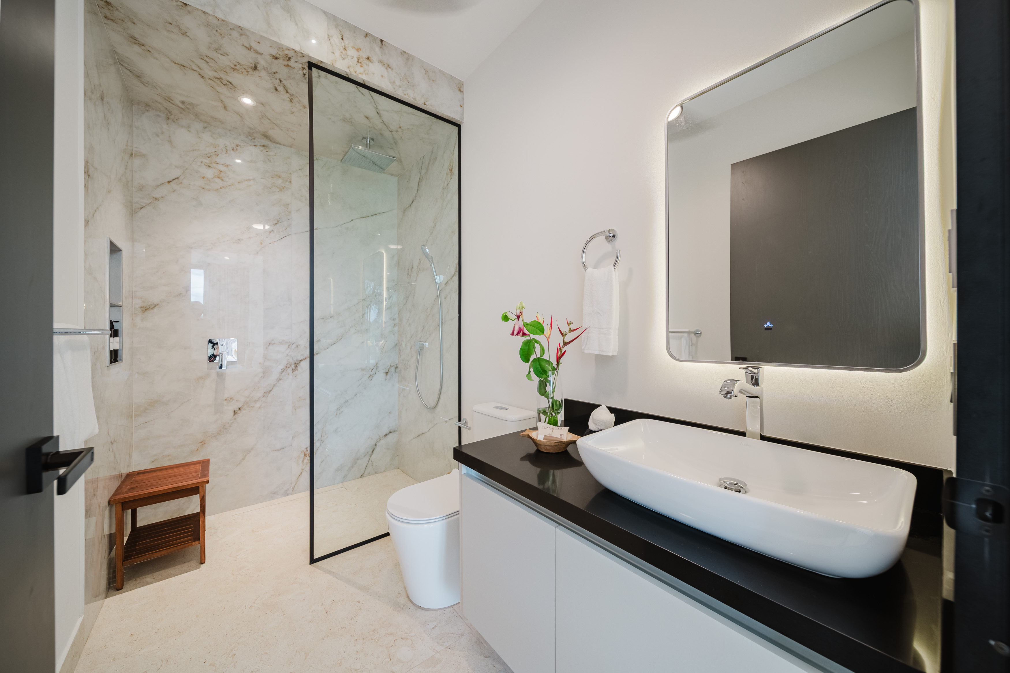This modern bathroom features a spacious walk-in shower with marble-look tiling, a glass enclosure, and an integrated wooden bench, alongside a sleek vanity with a vessel sink and a backlit mirror.