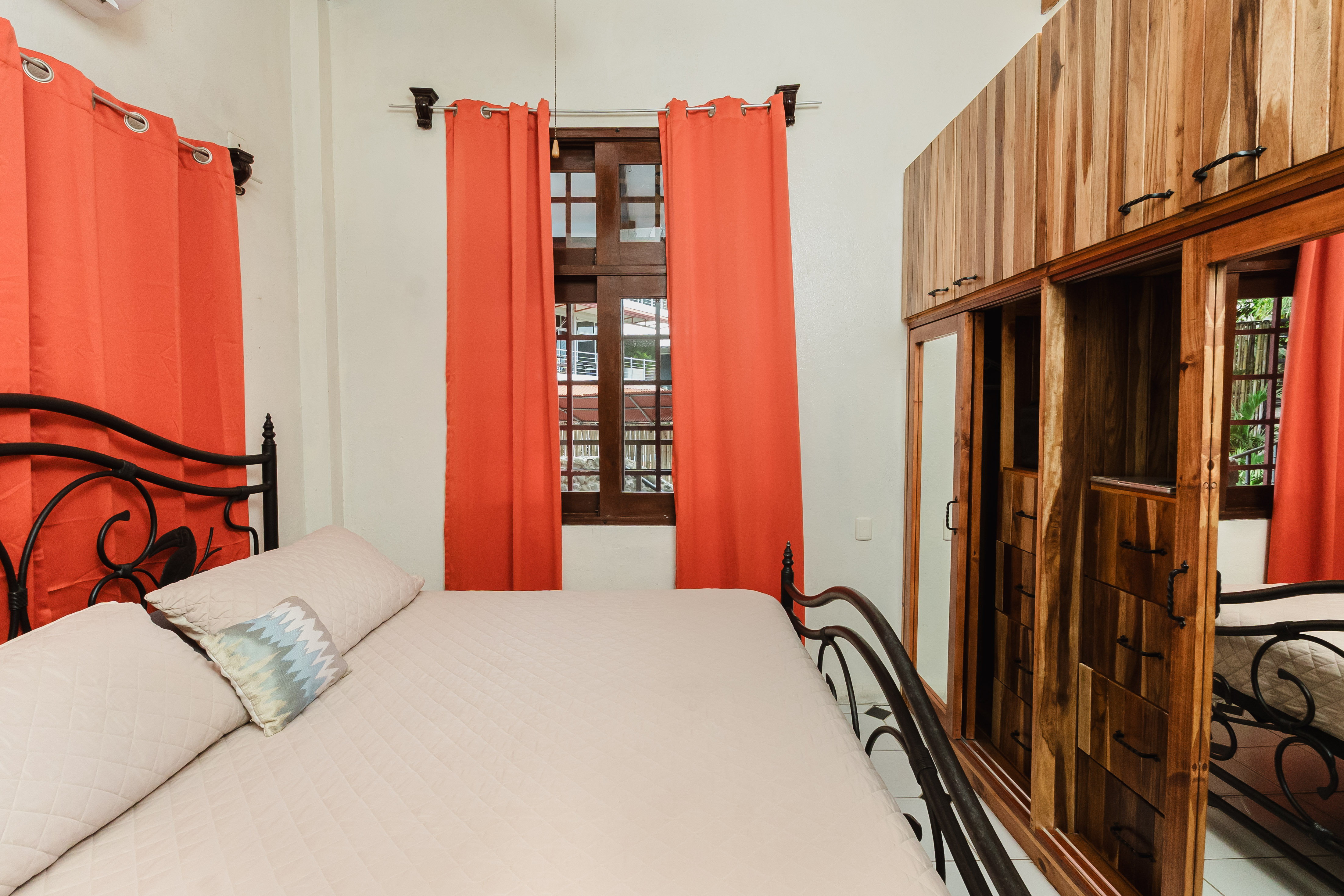 A bright bedroom combines a wrought-iron bed, natural light from wood-framed windows draped with vibrant orange curtains, and rich wooden cabinetry.