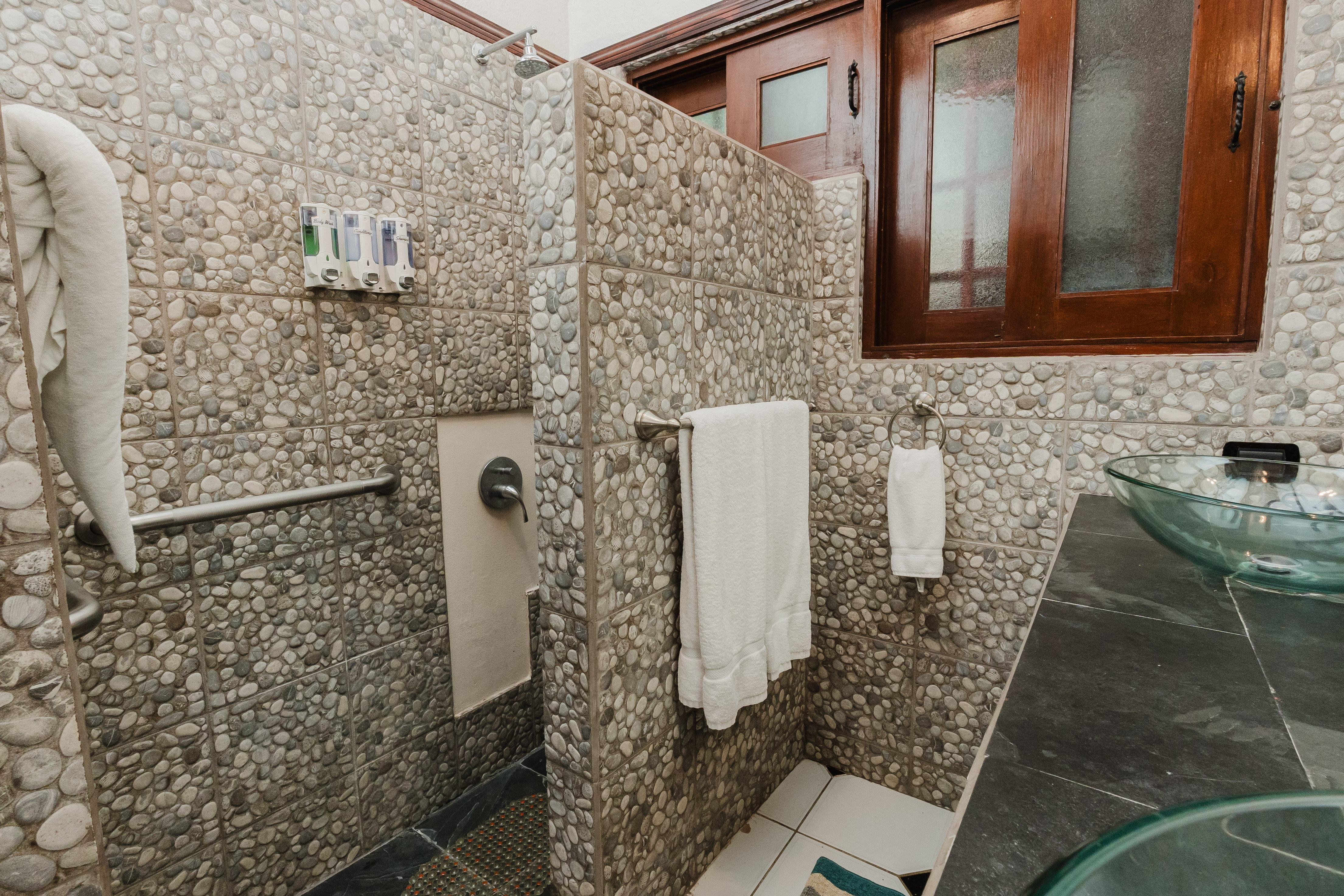 This bathroom features natural pebble-tiled walls, a rain shower, a glass vessel sink, and a privacy window, creating a serene, earthy atmosphere.