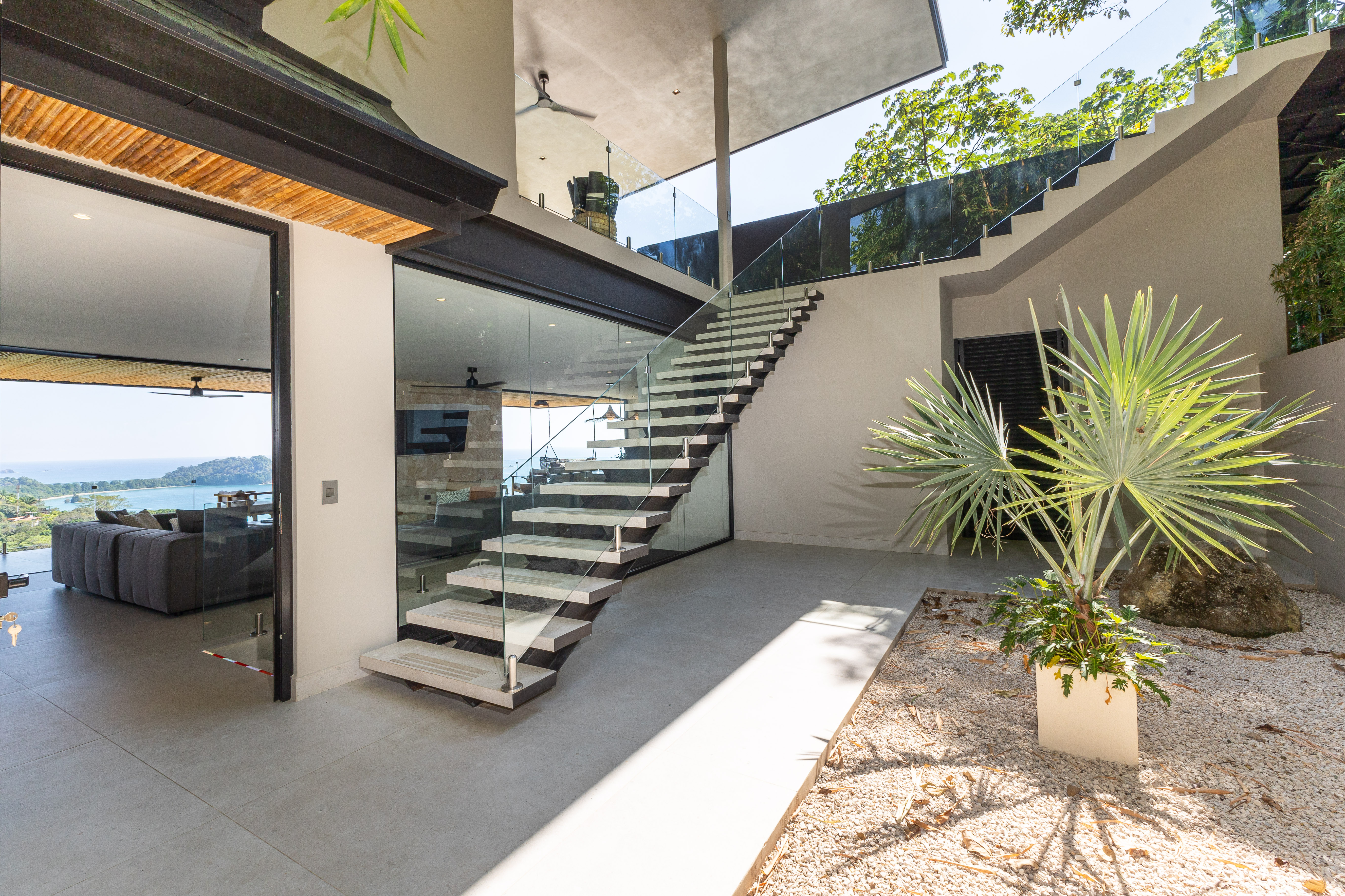 This bright, open entry features a floating staircase with glass railings, connecting to an outdoor garden and an indoor living area with floor-to-ceiling glass framing ocean and jungle views.