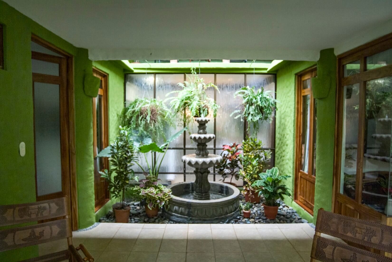 An indoor atrium with vibrant green walls features a multi-tiered fountain surrounded by tropical plants, illuminated by soft light from a textured glass window.