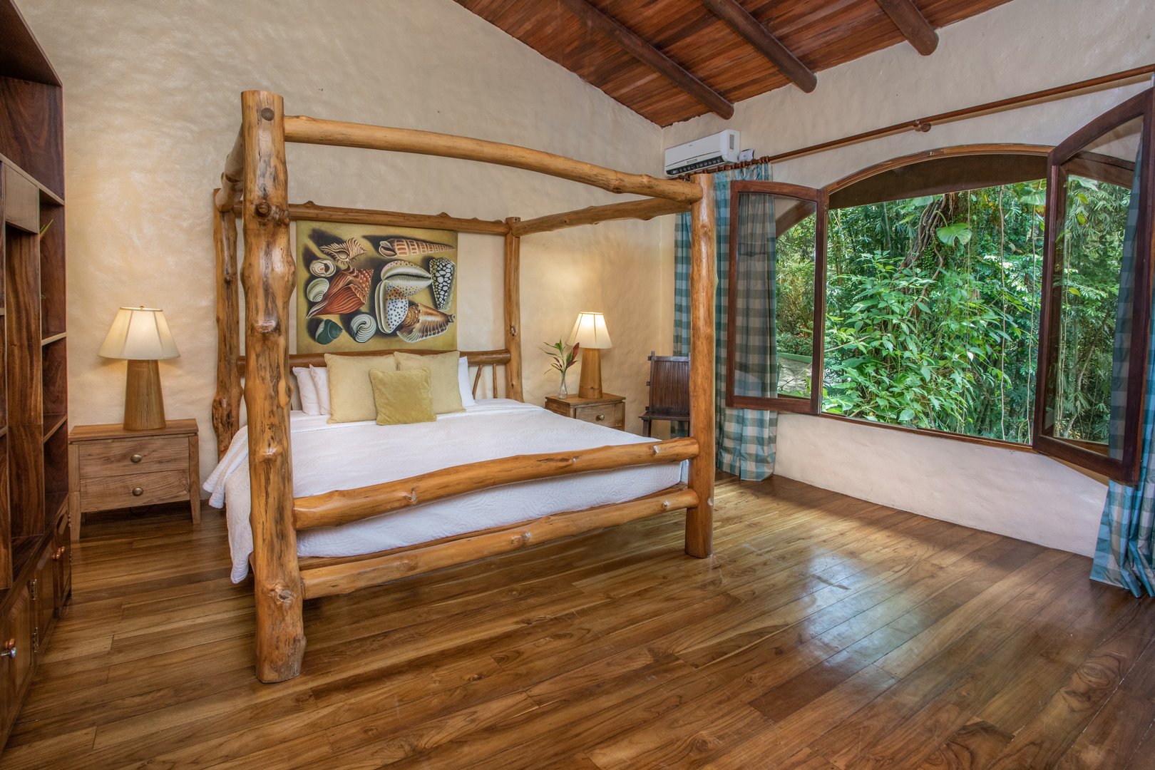 A bedroom with a rustic four-poster bed, exposed wood beam ceiling, hardwood floors, and a large open window providing direct views into a lush jungle.