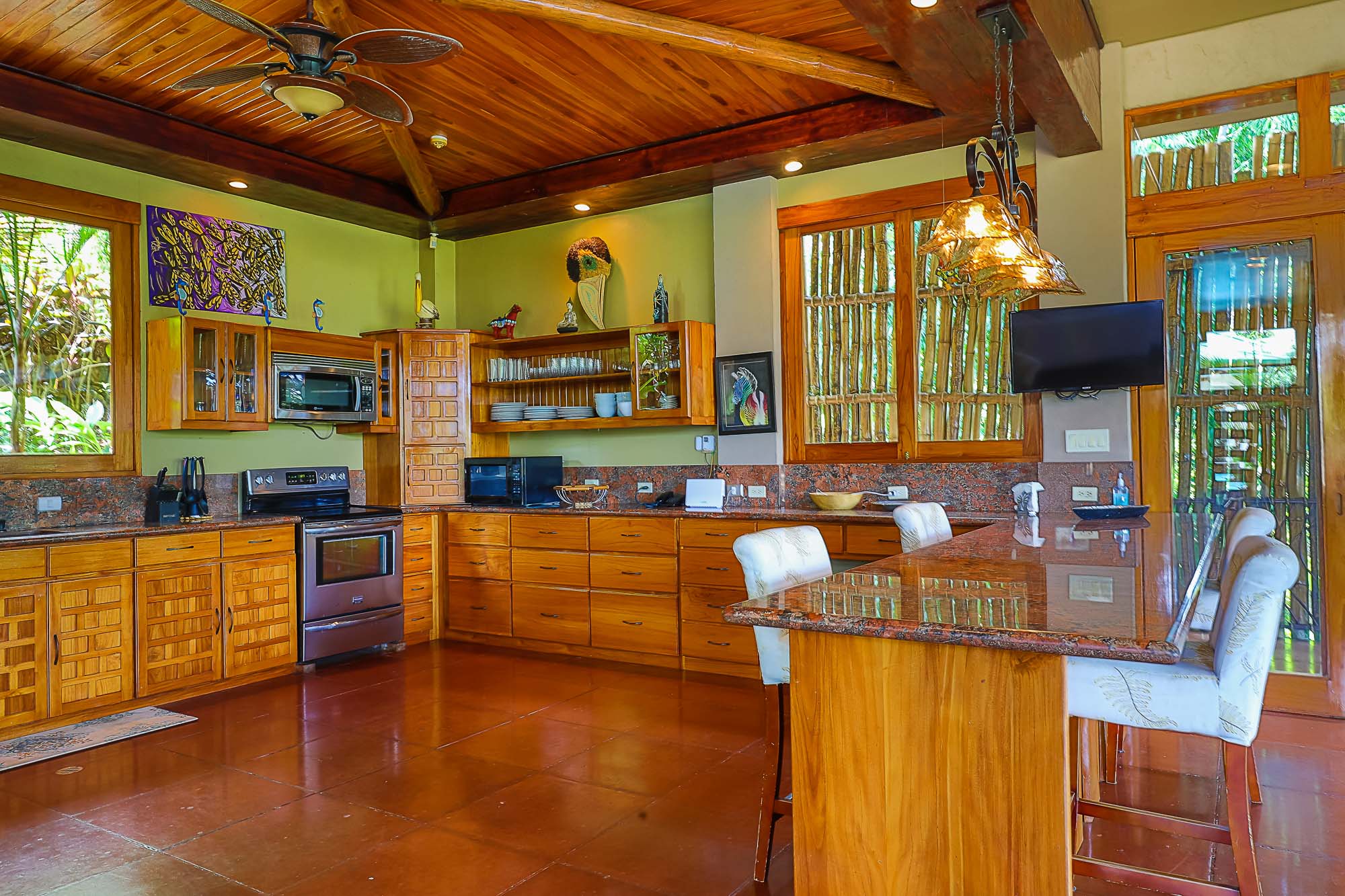 This kitchen, featuring extensive wooden cabinetry, granite countertops, and a large island, provides tropical views through wooden-framed windows and is capped by a high wood-paneled ceiling, offering a warm and open feel.