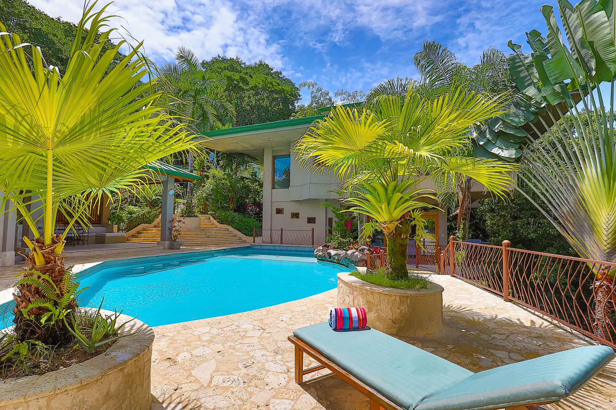 The outdoor pool area features a large swimming pool, a stone patio, and tropical foliage, with a contemporary multi-level villa visible against a backdrop of dense rainforest.