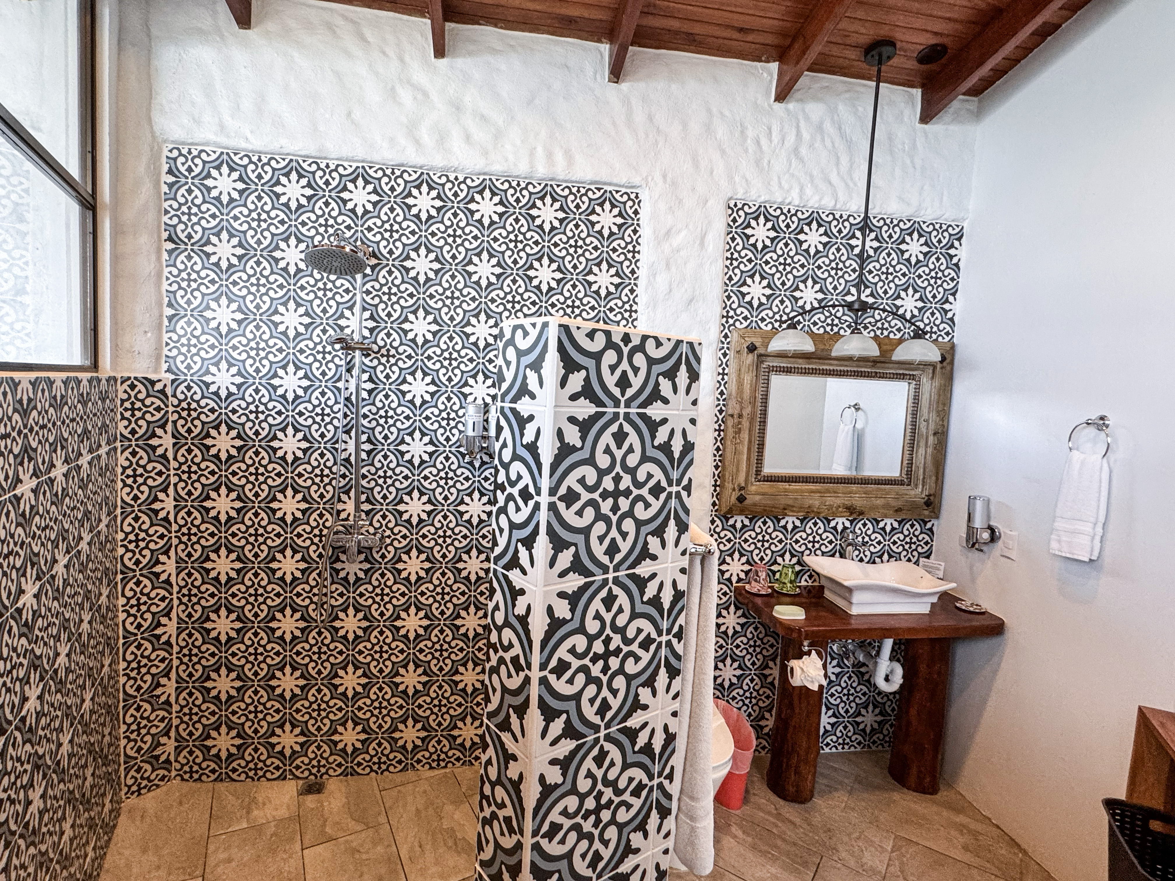 This bathroom features extensive patterned tile work on the walls and a central partition, alongside a rustic wooden vanity with a vessel sink and an exposed beam ceiling, brightened by natural light.