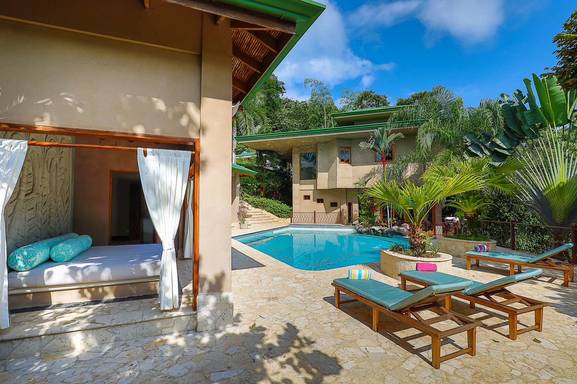 The sun-drenched outdoor area showcases a clear blue swimming pool, a stone patio with multiple sun loungers, and a shaded daybed pavilion, all framed by lush tropical foliage and the villa's tiered architecture.
