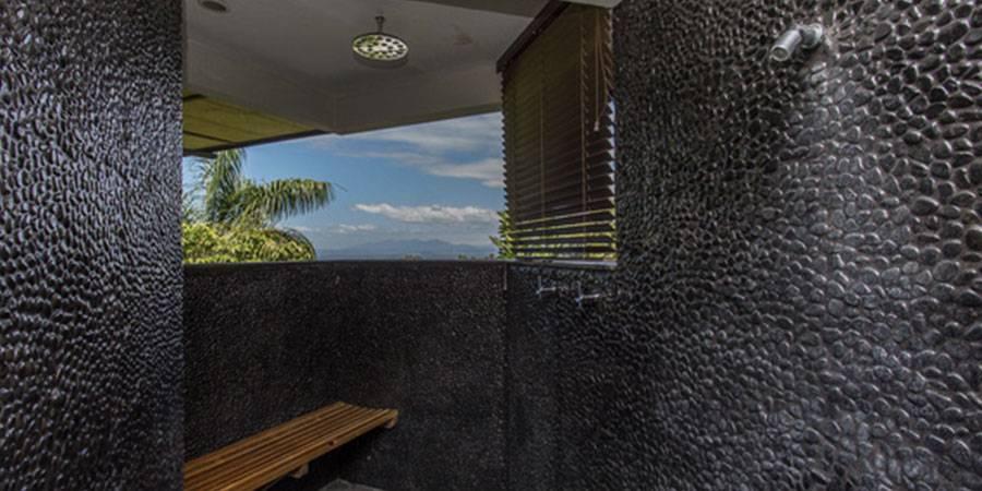 An open-air shower with dark pebble-textured walls and a wooden bench offers expansive views of tropical foliage and distant mountains.