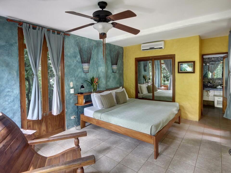 A bright bedroom suite with a wooden bed and colorful textured walls opens to a jungle view through large glass doors, revealing an ensuite bathroom.