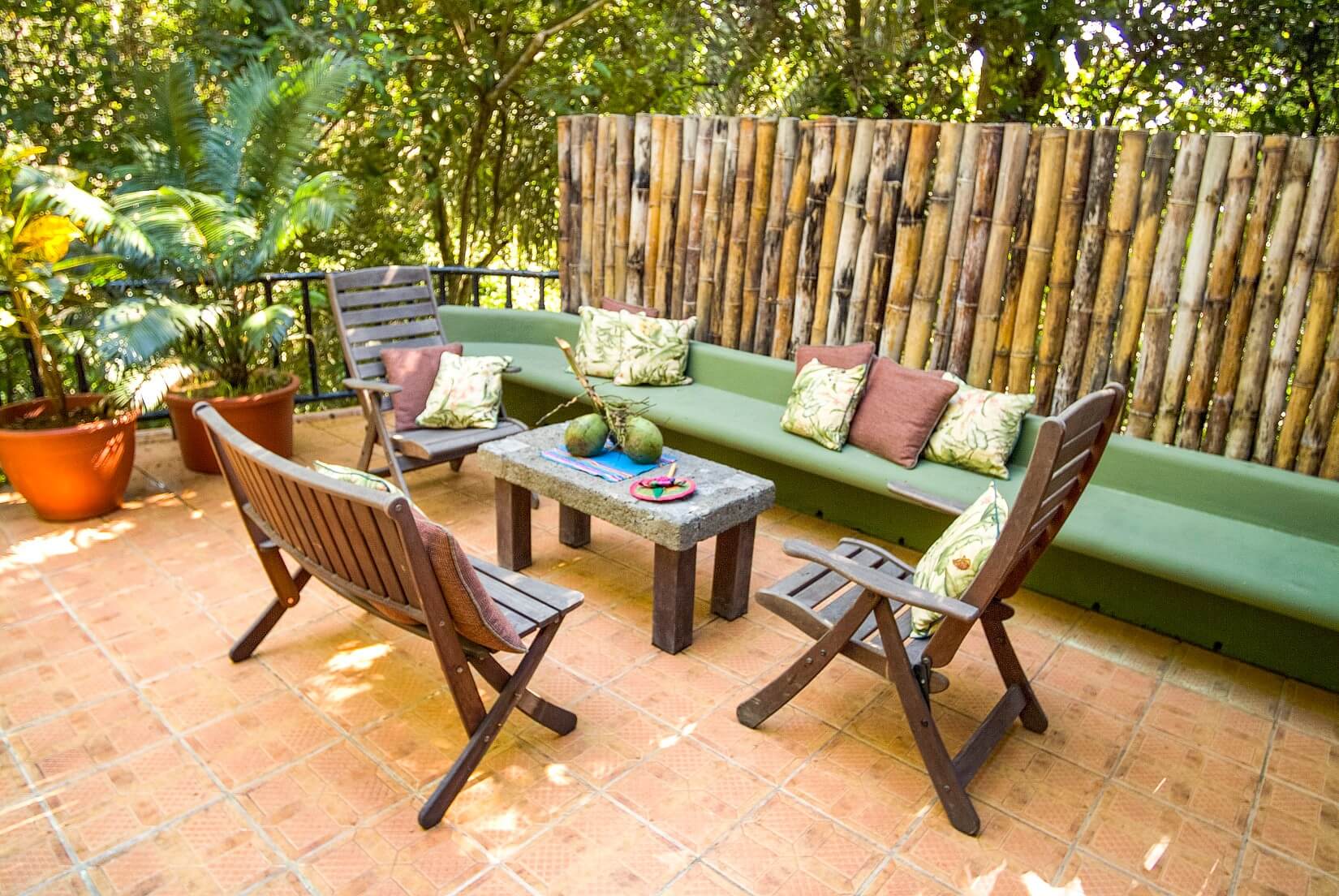 A secluded outdoor terrace offers a built-in green sofa and chairs on tiled flooring, enveloped by lush tropical plants and a bamboo privacy screen.