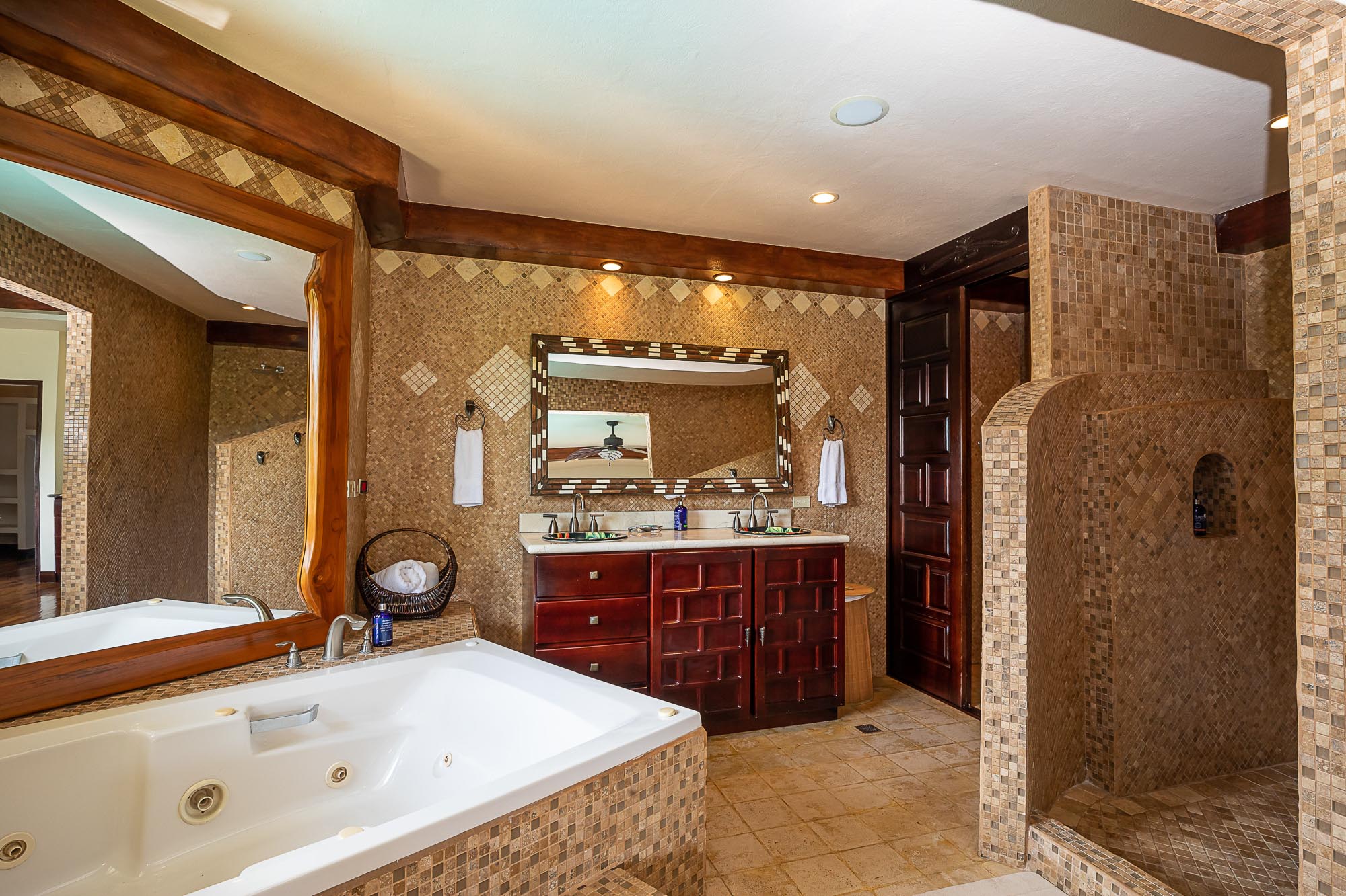 A spacious bathroom designed with warm mosaic tiling and dark wood accents features a jetted tub, a walk-in shower, and a dual vanity with a patterned mirror.