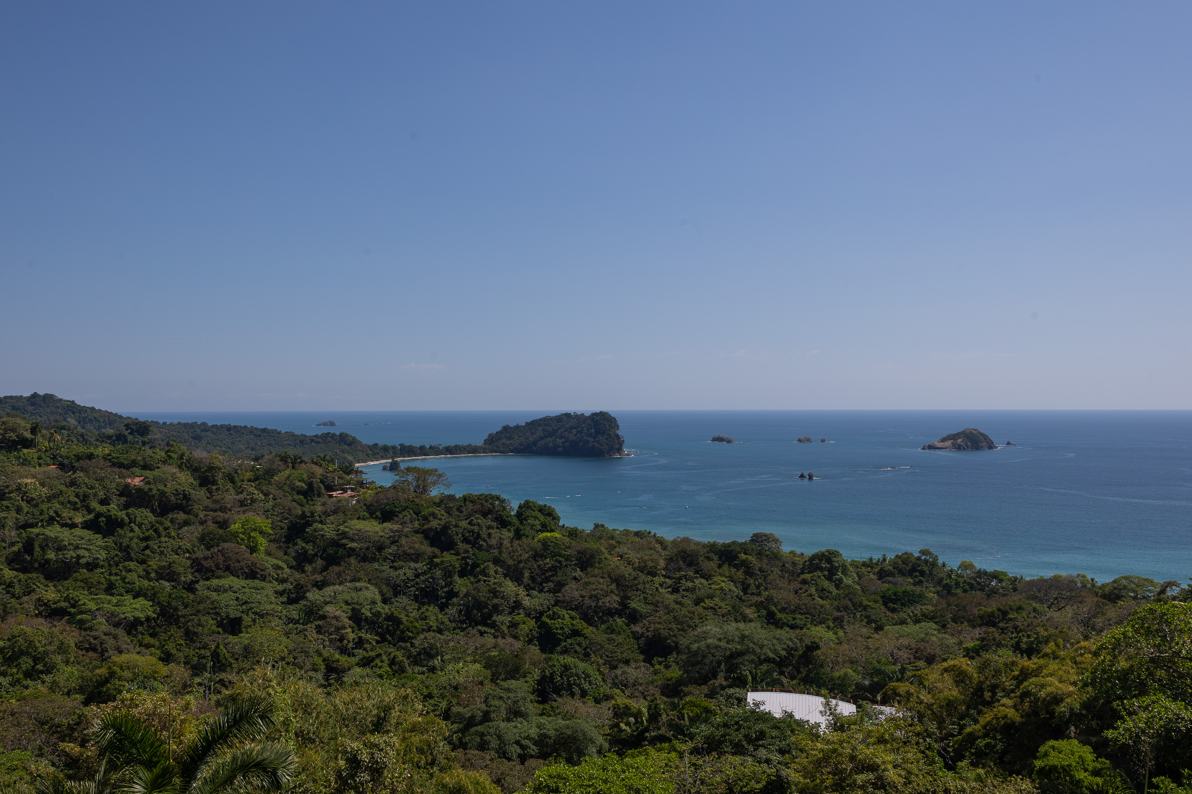 A clear blue sky illuminates a vast ocean view, showcasing forested islands and a curving coastline framed by dense tropical jungle.